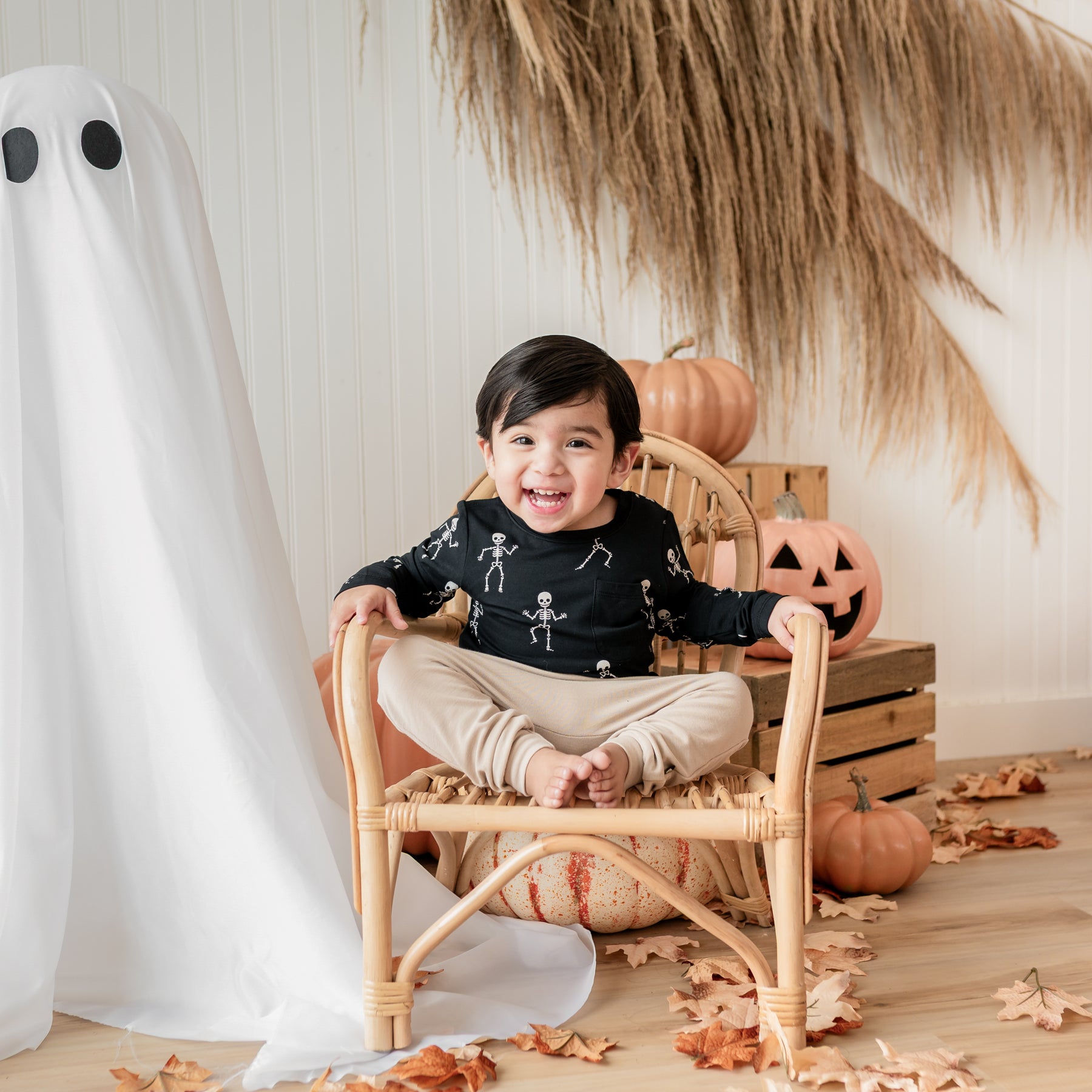 Smiling toddler sitting on a wicker chair wearing the Long Sleeve Toddler Crew Neck Tee in Skeleton in front of wooden crates, pumpkins, leaves and a sheet ghost