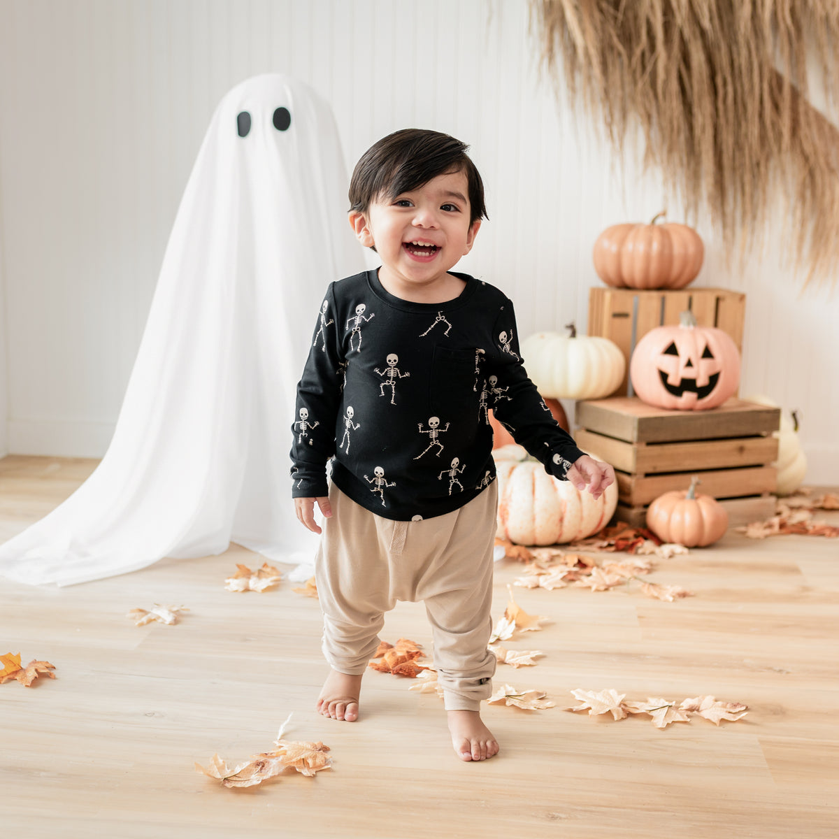 Young boy walking in front of a fall backdrop of a sheet ghost, pumpkins, wooden crates and leaves wearing the Long Sleeve Toddler Crew Neck Tee in Skeleton and light colored bamboo jersey harem pant