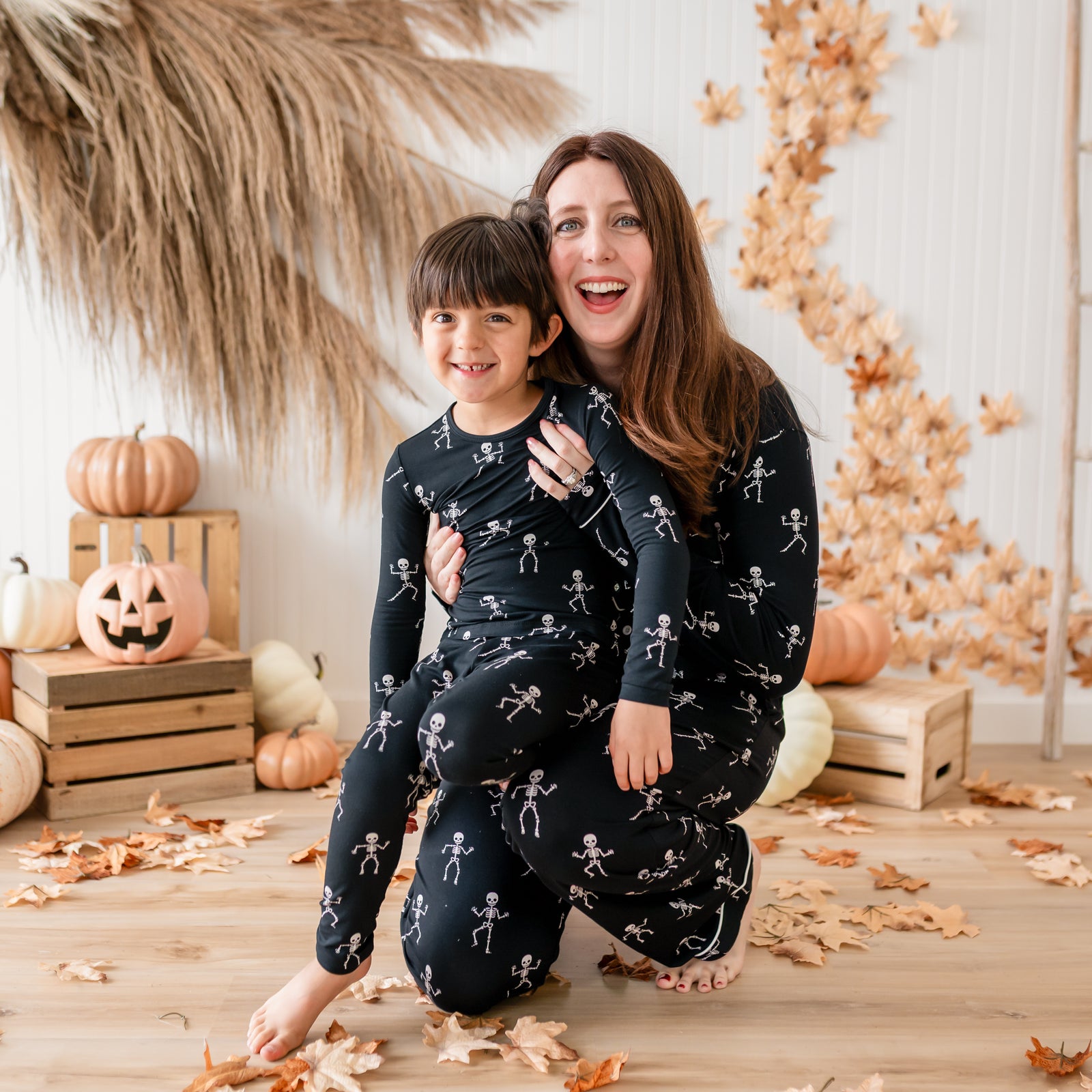 Mother and son matching in Skeleton. Mom kneeling on the ground wearing the Women's Long Sleeve Pajama Set holding her son on her lap who is in the long sleeve toddler pajamas. Both are in front of a fall backdrop of pumpkins, wooden crates and leaves