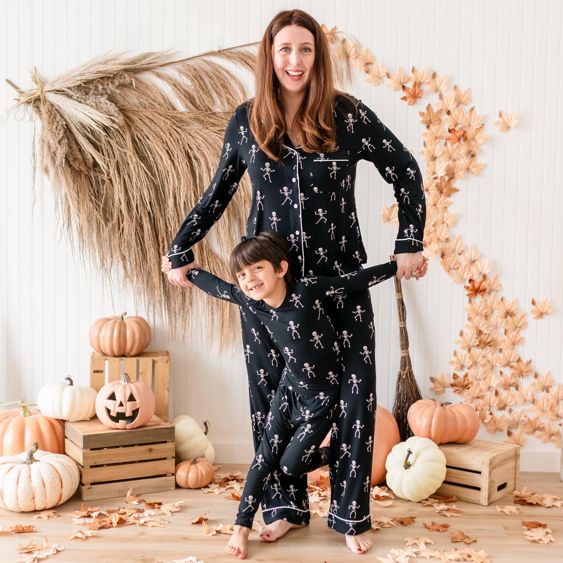 Mom and son matching in Skeleton. Mother standing behind son holding his hands in the air wearing the Women's Long Sleeve Pajamas and son wearing the toddler pajamas. Both are standing in front of a fall backdrop