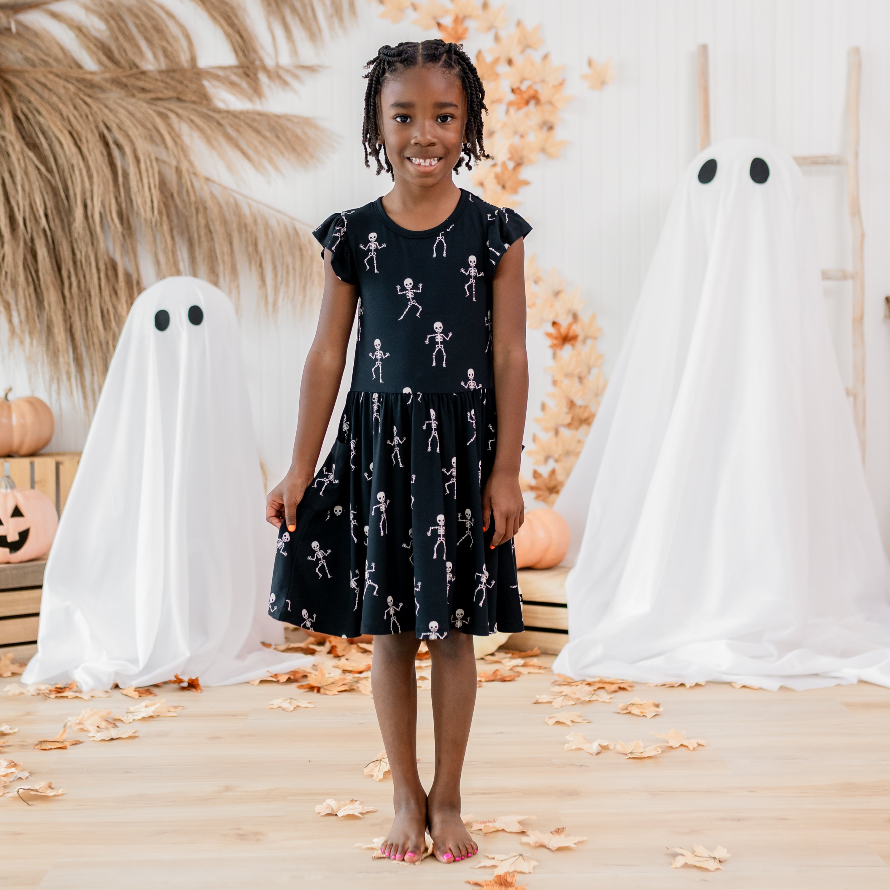 Young girl model standing between two sheet ghosts wearing the Pocket Dress in Skeleton holding the side of the dress out slightly