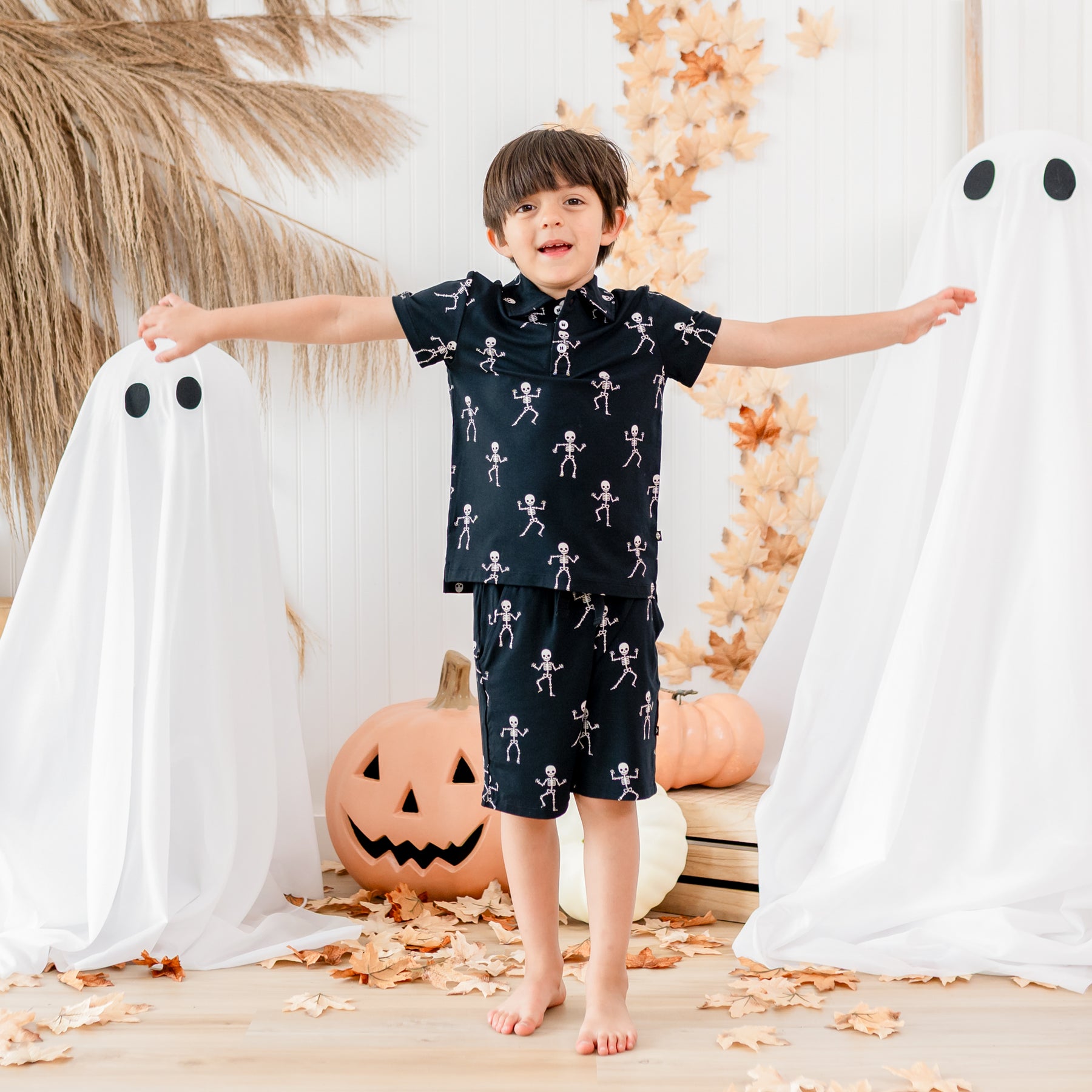 Boy modeling the Toddler Short Sleeve Polo in Skeleton with his arms out. Standing in front of sheeted ghosts, pumpkins, wooden crates and leaves
