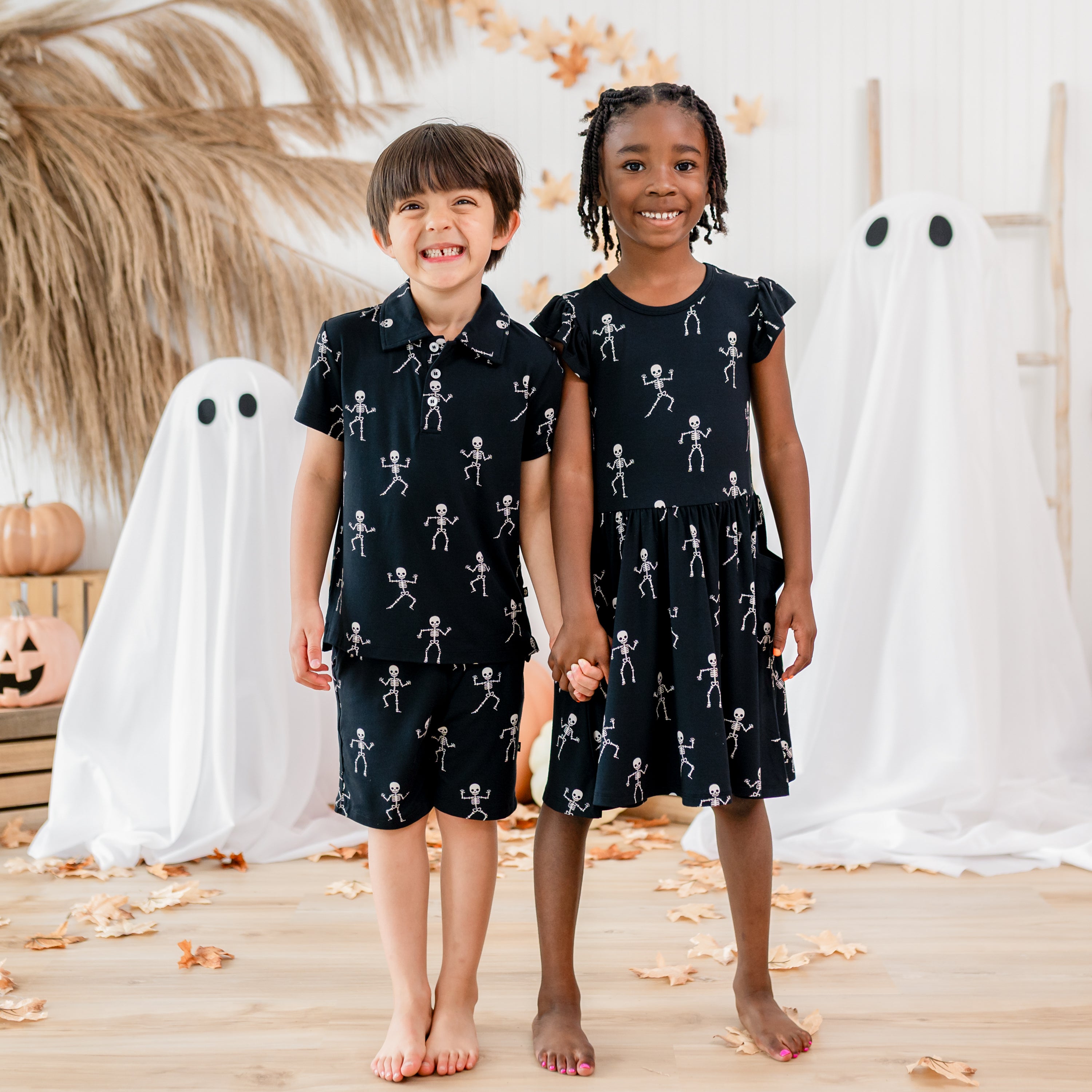 Two smiling children holding hands wearing various Skeleton items in front of a fall back drop. Items include a pocket dress, short sleeve polo shirt and drawstring shorts