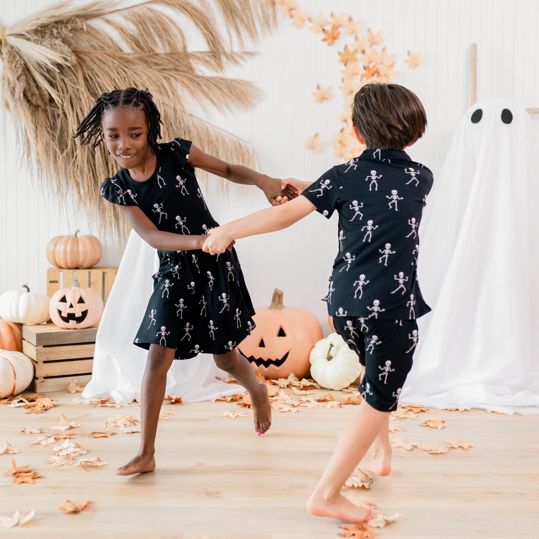 Two children spinning around wearing the Skeleton print in front of a fall backdrop. Pictured is the Drawstring Short, short sleeve toddler polo and pocket dress