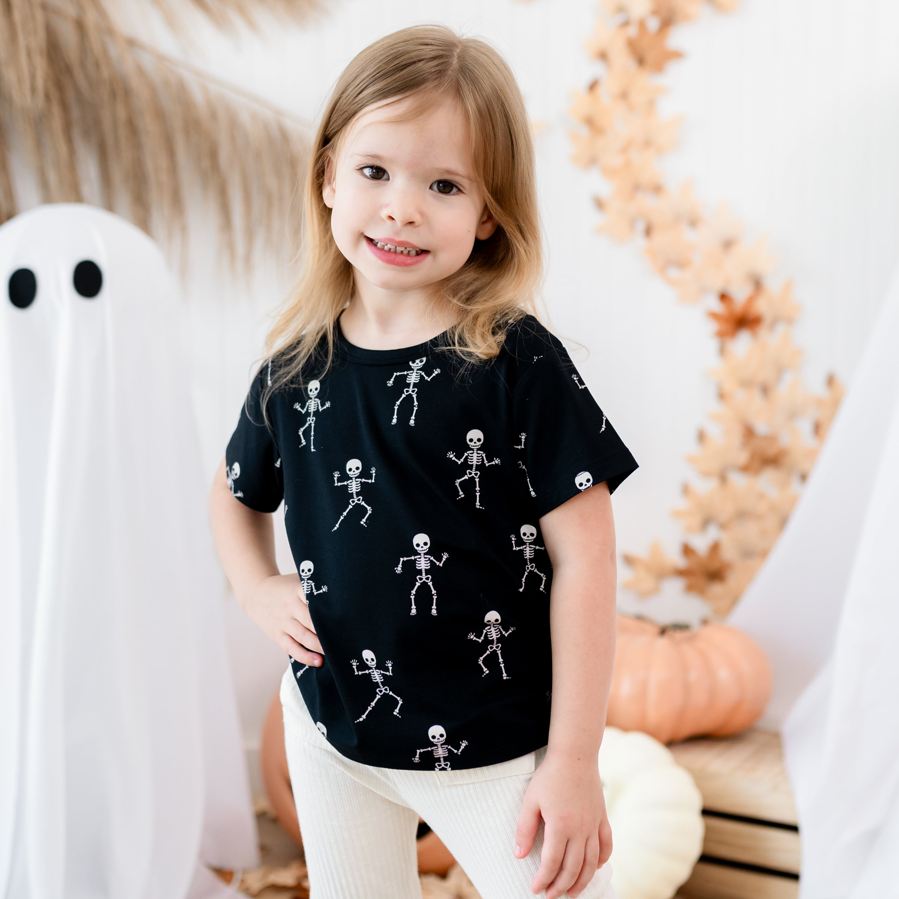 Smiling young girl with hand on her hip wearing the Toddler Basic Tee in Skeleton in front of sheet ghosts, leaves, pumpkins and wooden crates
