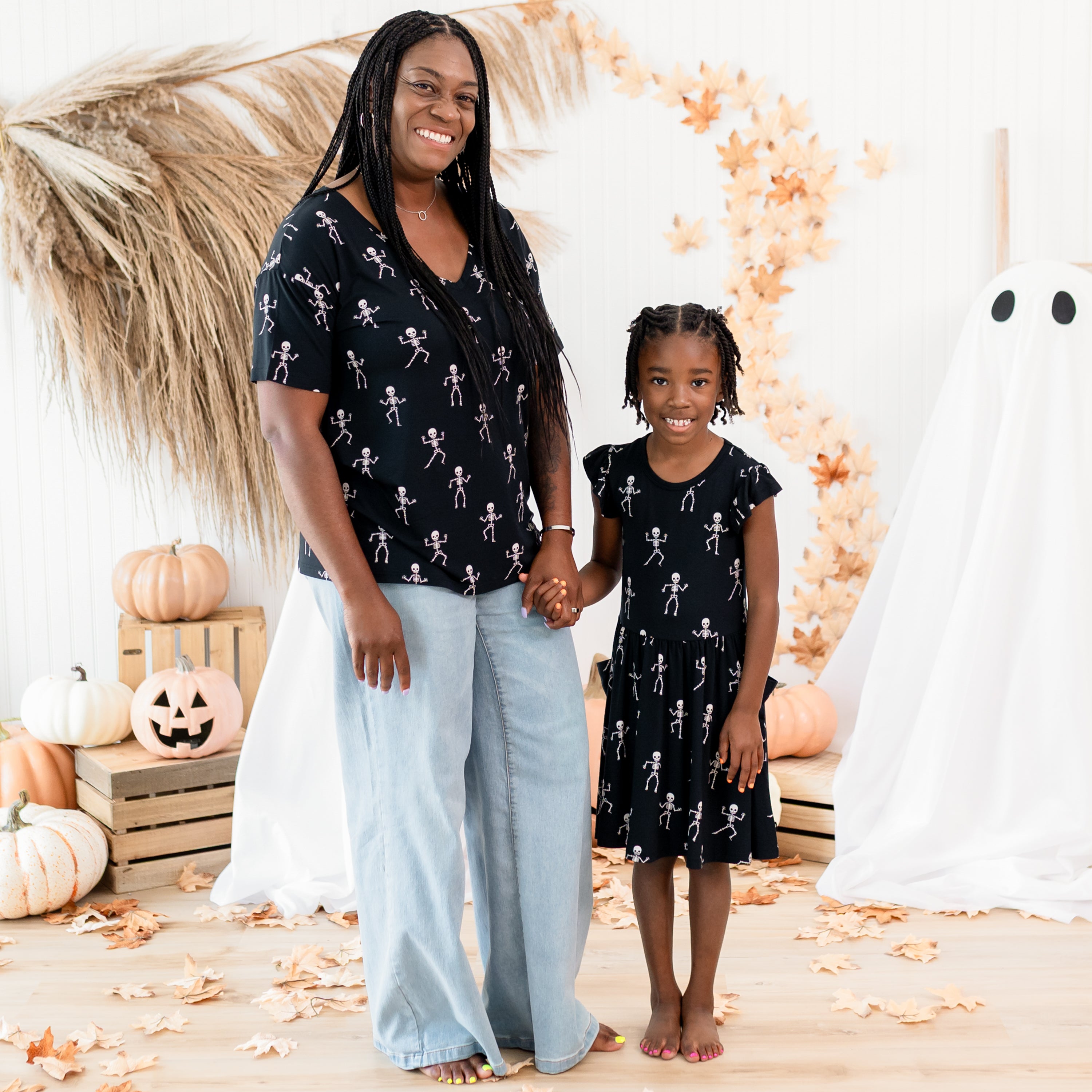 Mother and daughter matching in Skeleton. Mother wearing the Women's Relaxed Fit V-Neck and light wash jeans holding daughters hand wearing the pocket dress. Both standing in front of  fall inspired backdrop consisting of sheet ghosts, pumpkins, wooden crates and leaves