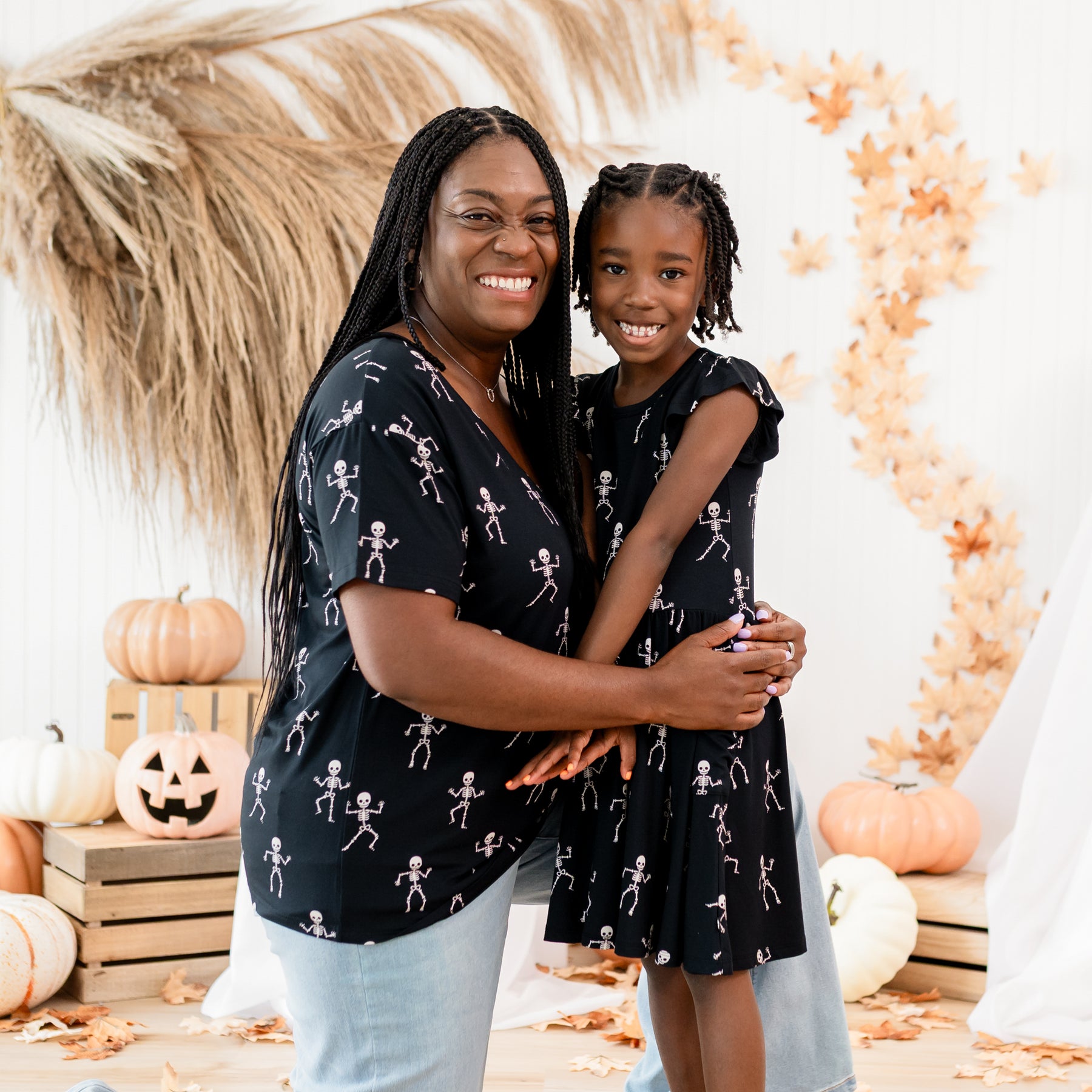 Mother and daughter matching in Skeleton. Mother is kneeling wearing the Women's Relaxed Fit V-Neck and jeans with her arms wrapped around her daughter who is wearing the pocket dress. Both are in front off a rustic fall inspired, halloween back drop