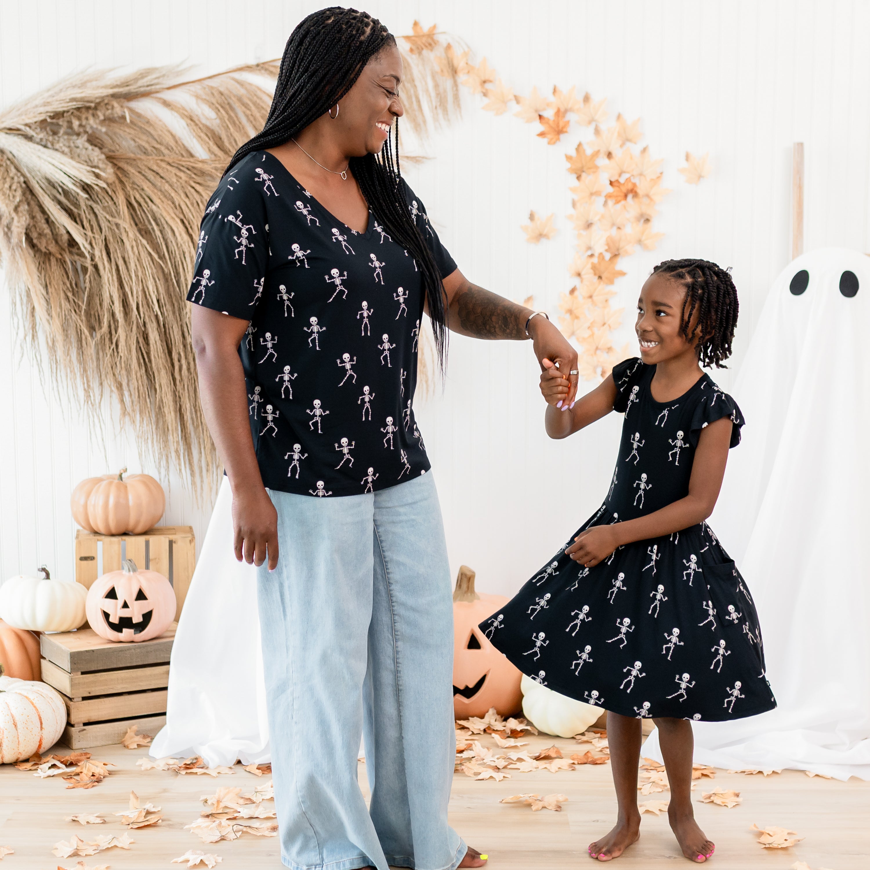 Mother and daughter matching in Skeleton. Mother wearing the women's v-neck is twirling her daughter who is wearing the pocket dress. Both are in front of a fall backdrop