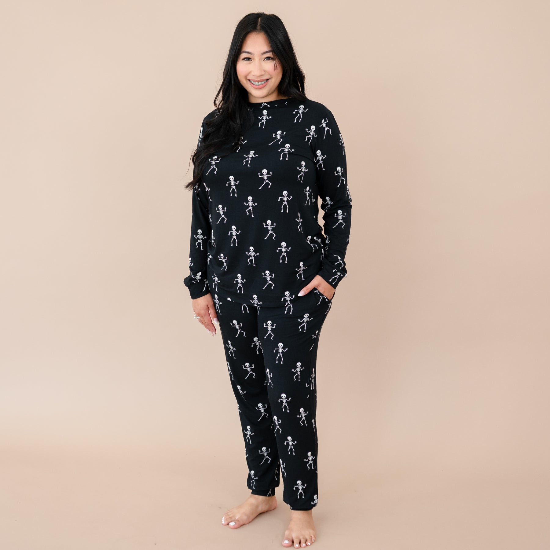 Smiling female wearing the Women's Jogger Pajama Set in Skeleton with one hand in the pants pocket in front of a taupe back drop