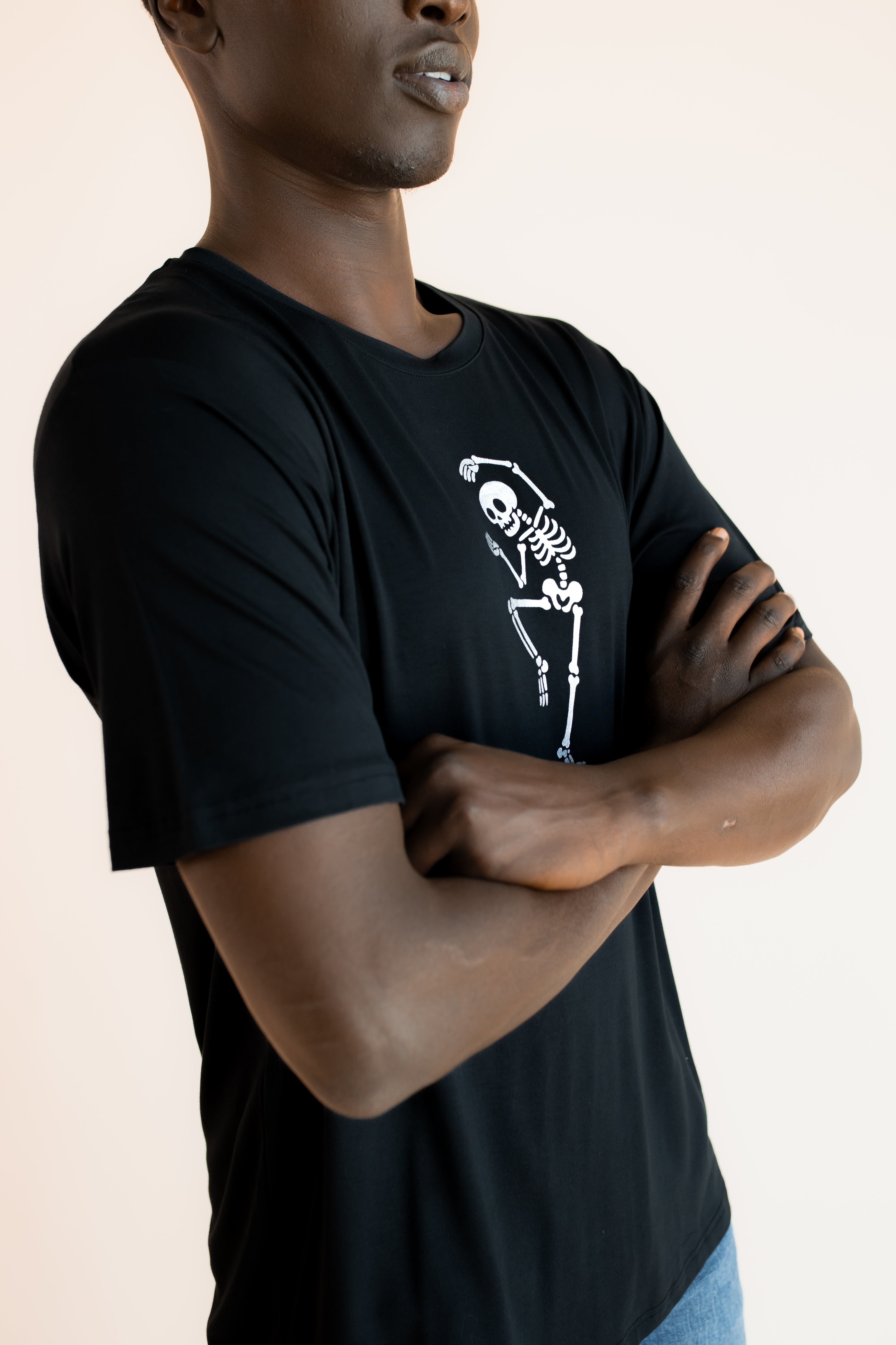 Side pose of Modal wearing Black Men's Crew Neck Tee with Skeleton print in center on a beige background