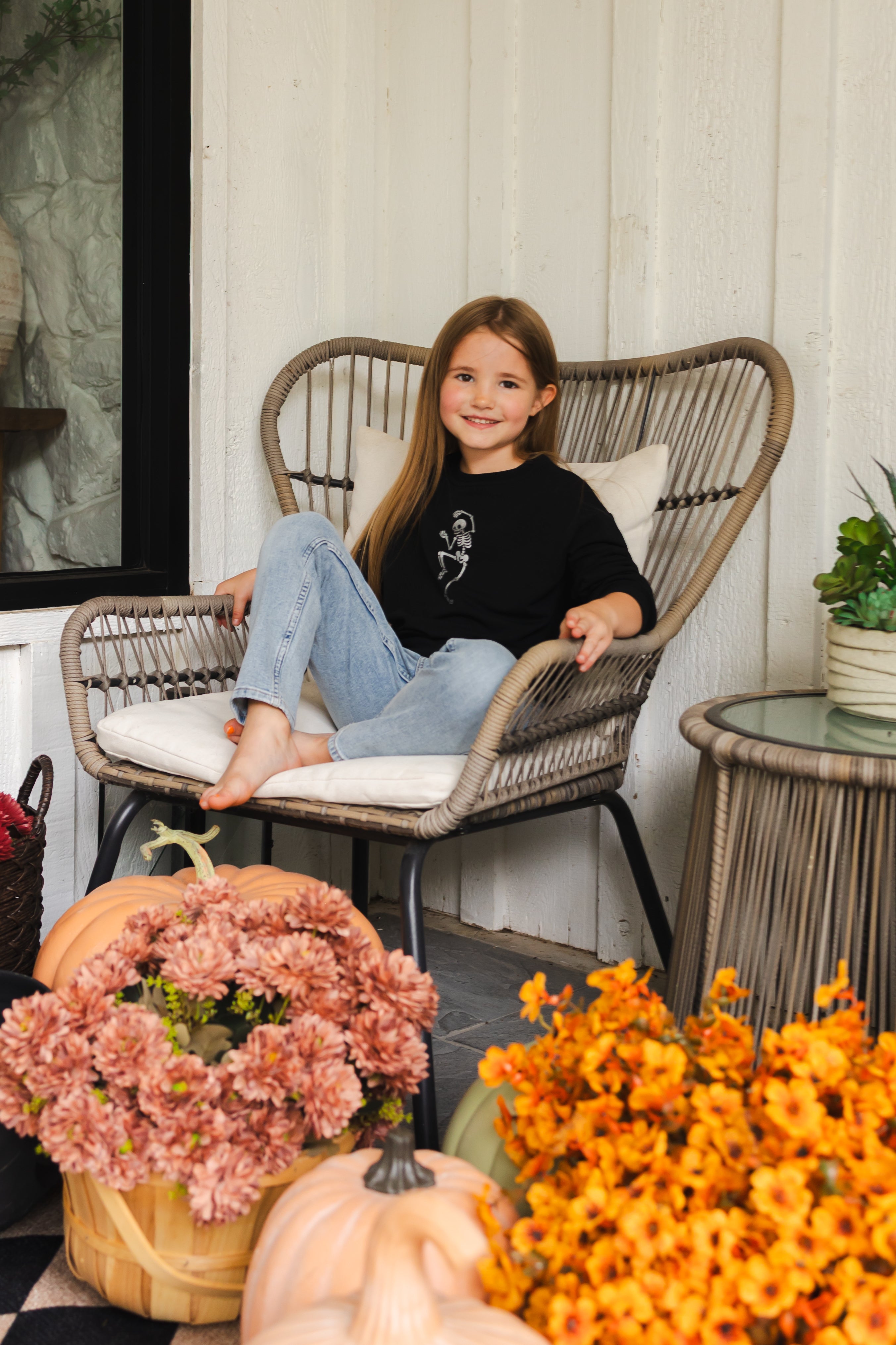 Girl wearing Black Sweatshirt with Skeleton print in the center sitting on a chair with flowers around