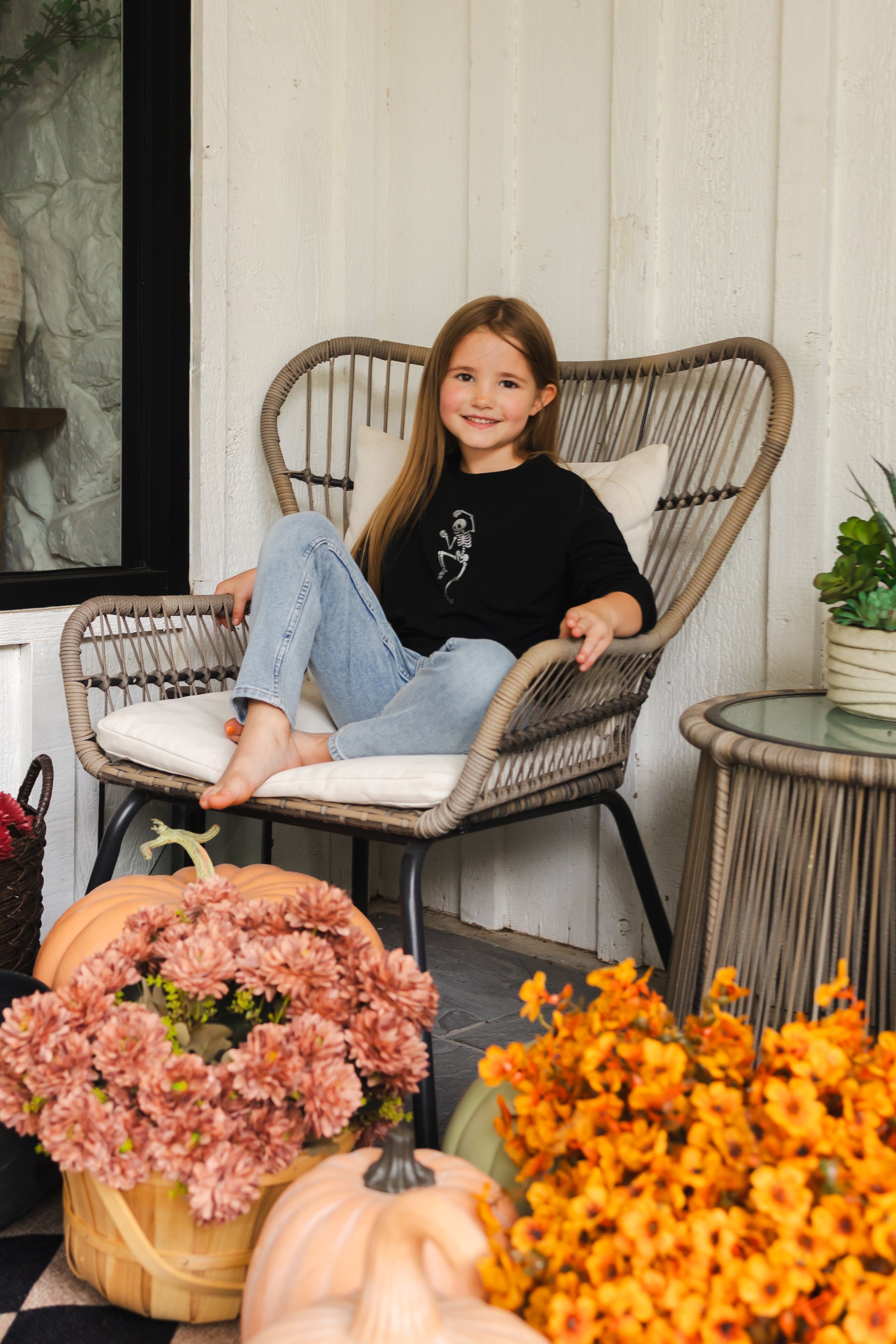 Girl wearing Black Sweatshirt with Skeleton print in the center sitting on a chair with flowers around