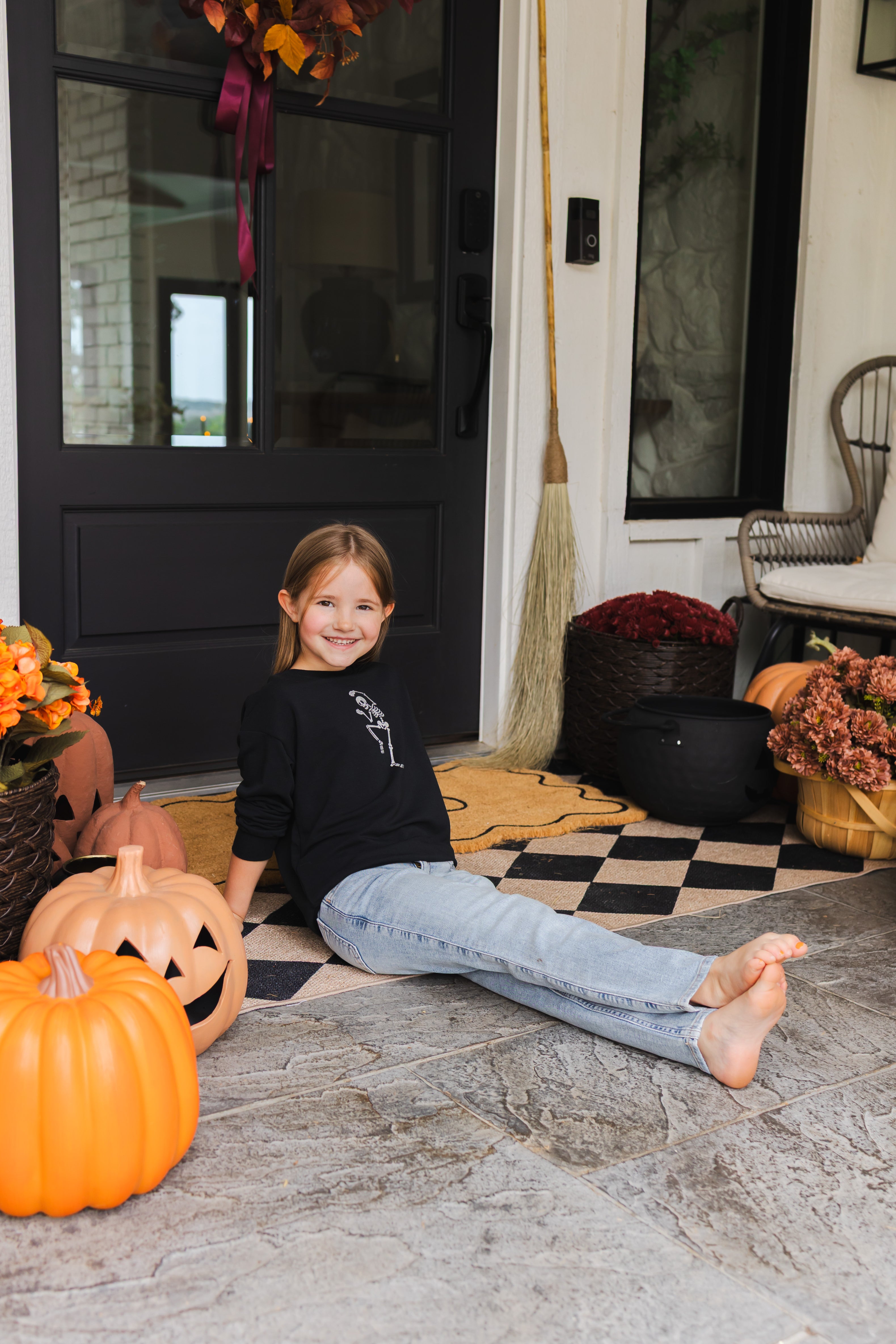 Girl smiling in Black Sweatshirt with Skeleton print in the center sitting outdoors with Halloween decorations