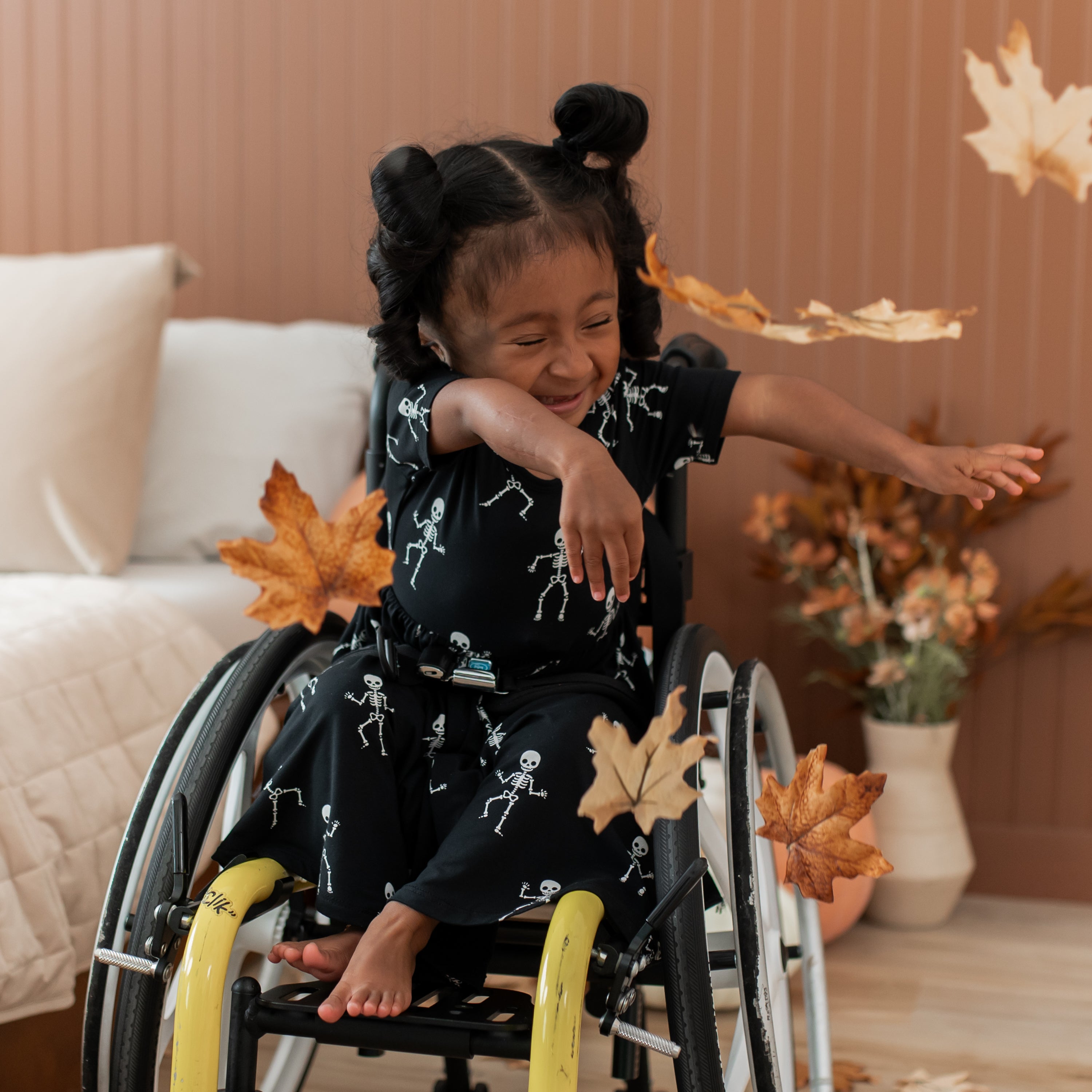 Child in a wheelchair with skeleton print black dress surrounded by autumn leaves indoors.