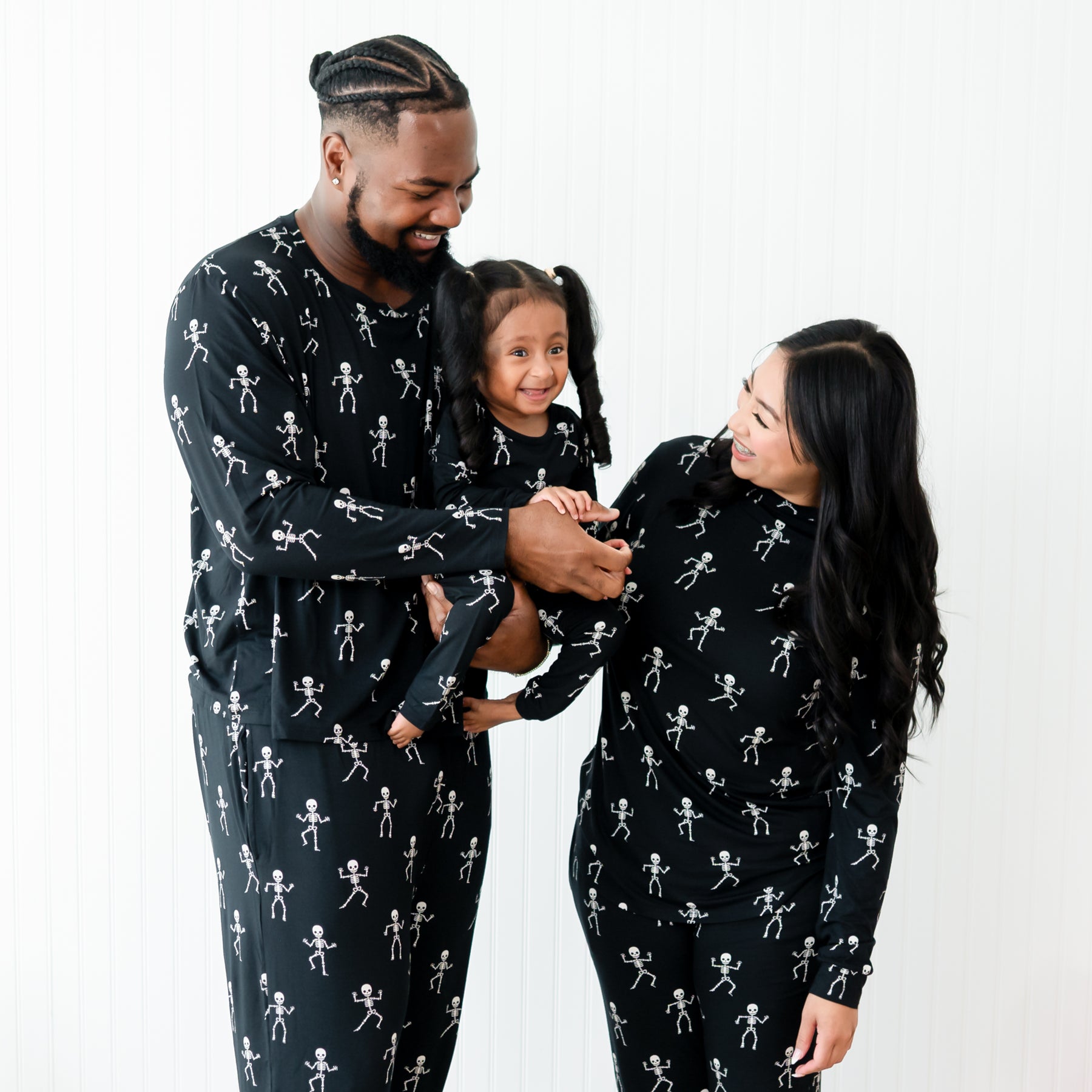 Family matching in Skeleton. Close up of the family, father is wearing the Men's Jogger Set holding his daughter who wearing toddler pajamas. The mother is standing beside them smiling looking at their daughter wearing the Women's jogger set