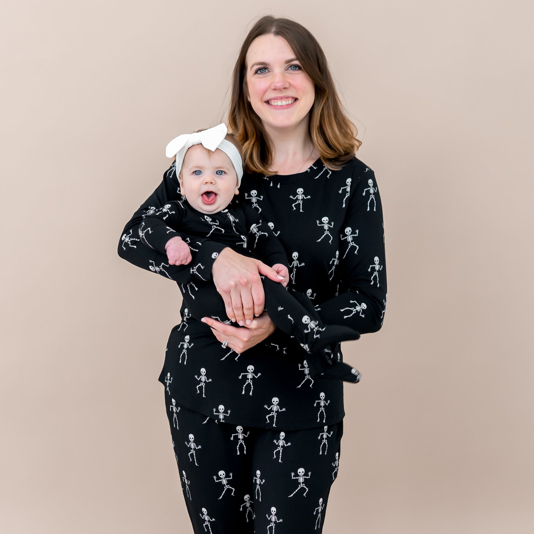 Mother and daughter matching in Skeleton. Smiling mother wearing the Women's Jogger Pajama Set holding her infant daughter who is wearing the zippered footie in front of a taupe backdrop