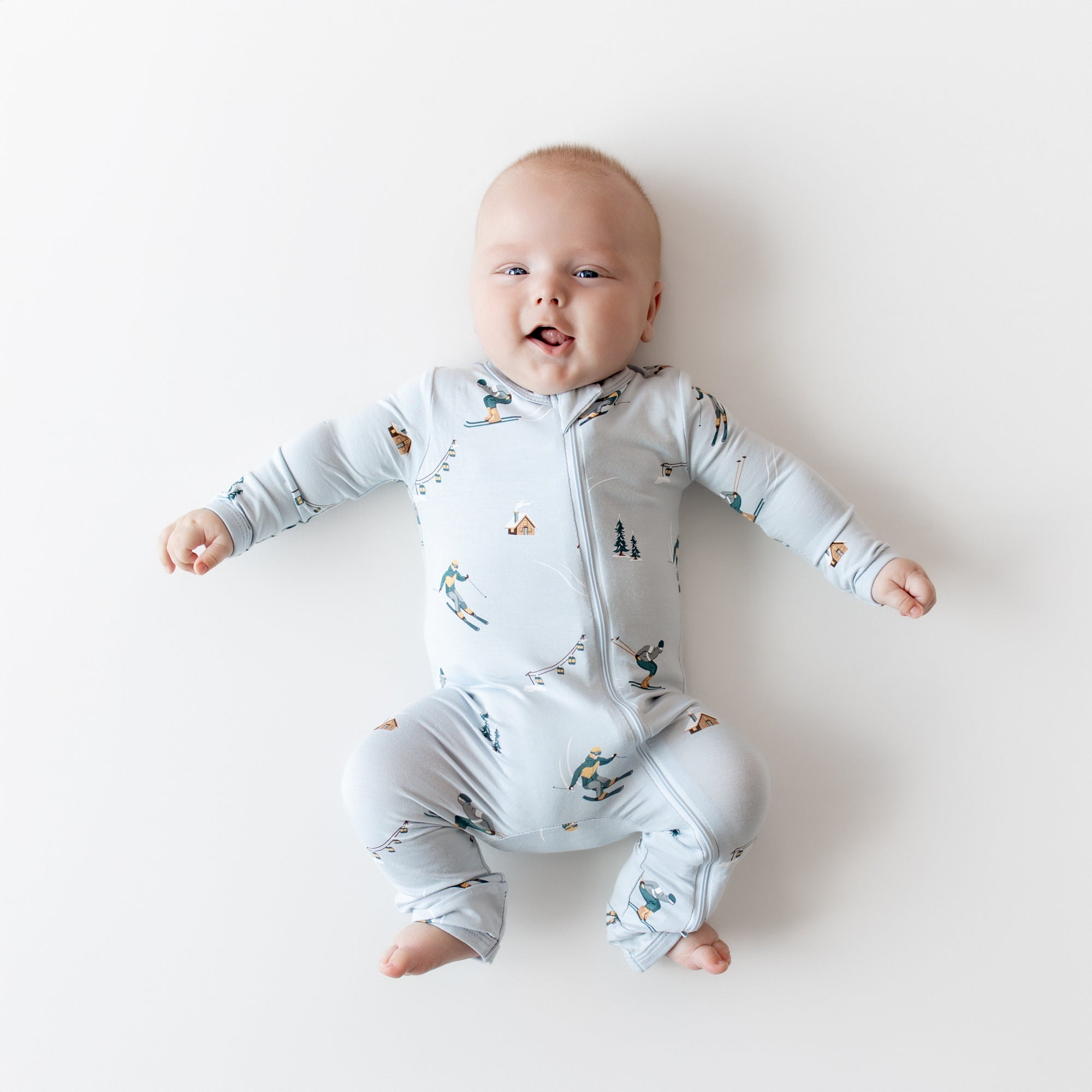 Smiling infant laying on a light colored surface wearing the Zippered Romper in Ski