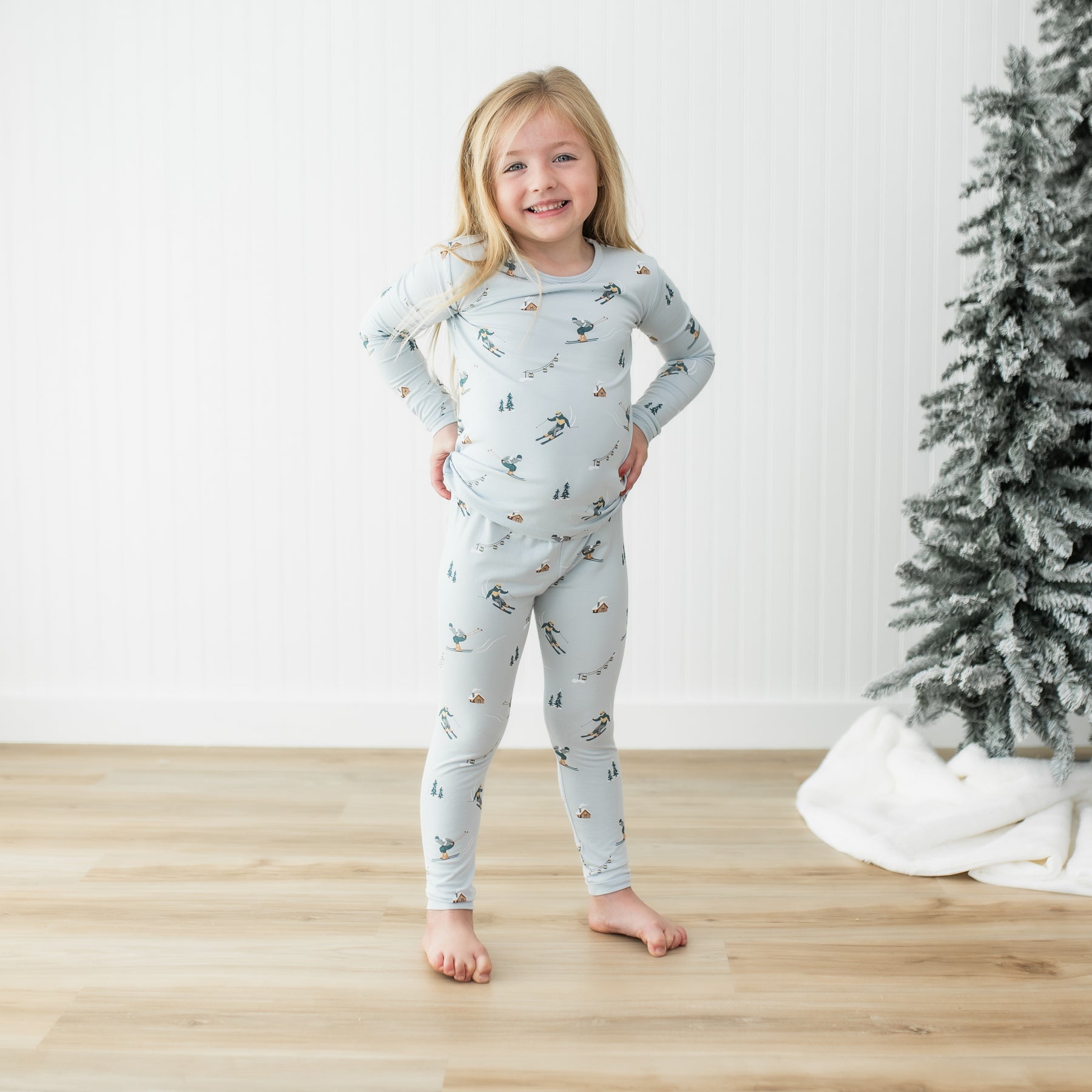 Young girl standing on a light wood floor wearing the Long Sleeve Pajamas in Ski with both hands on hips standing beside a frosted tree