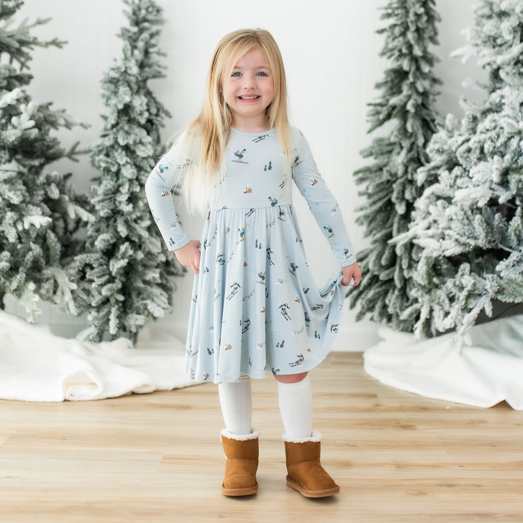 Smiling girl standing in front of frosted trees wearing the Long Sleeve Twirl Dress in Ski holding the hem of the twirl skirt