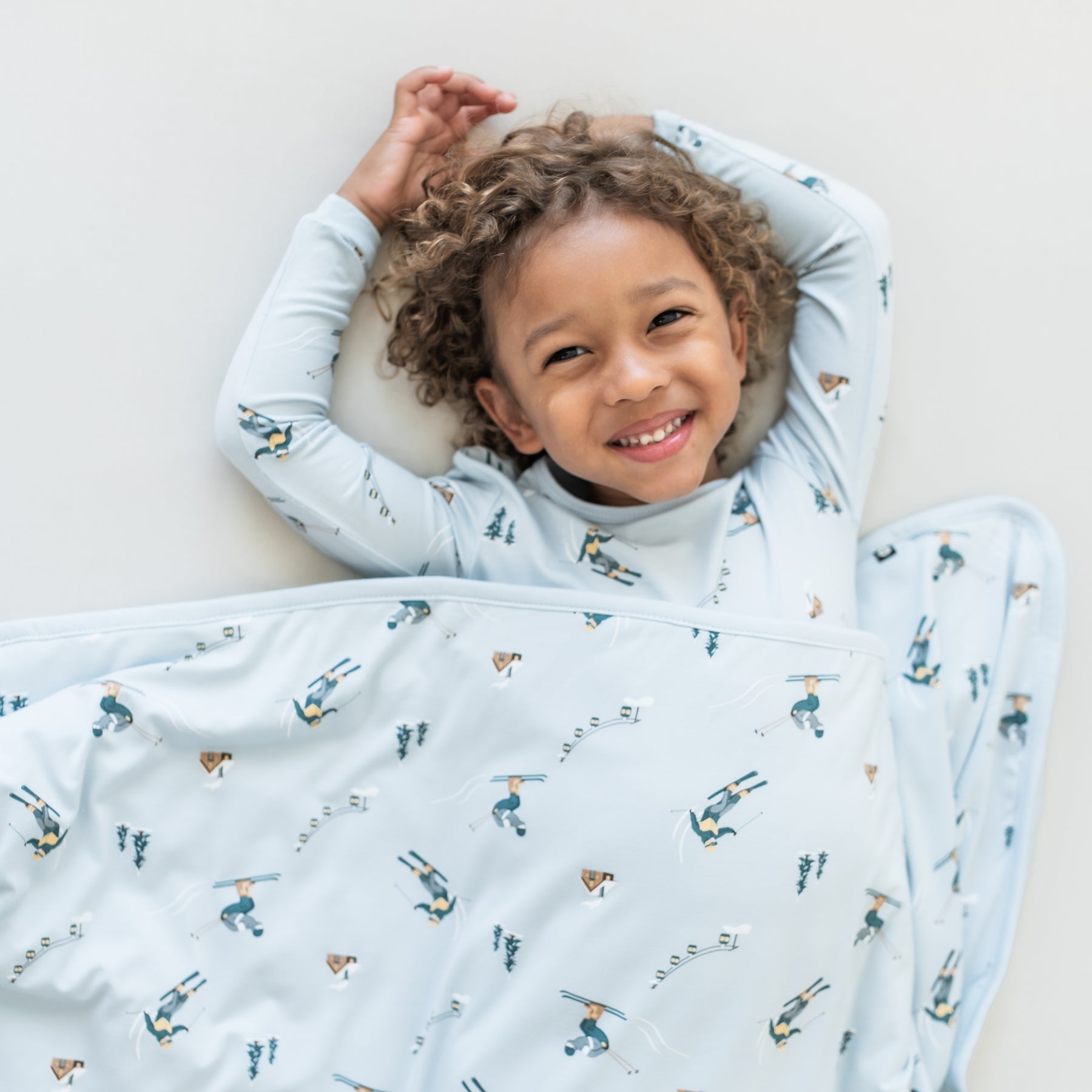 Young smiling child laying on a light colored surface wearing the Long Sleeve Pajamas in Ski with a matching blanket overtop of them