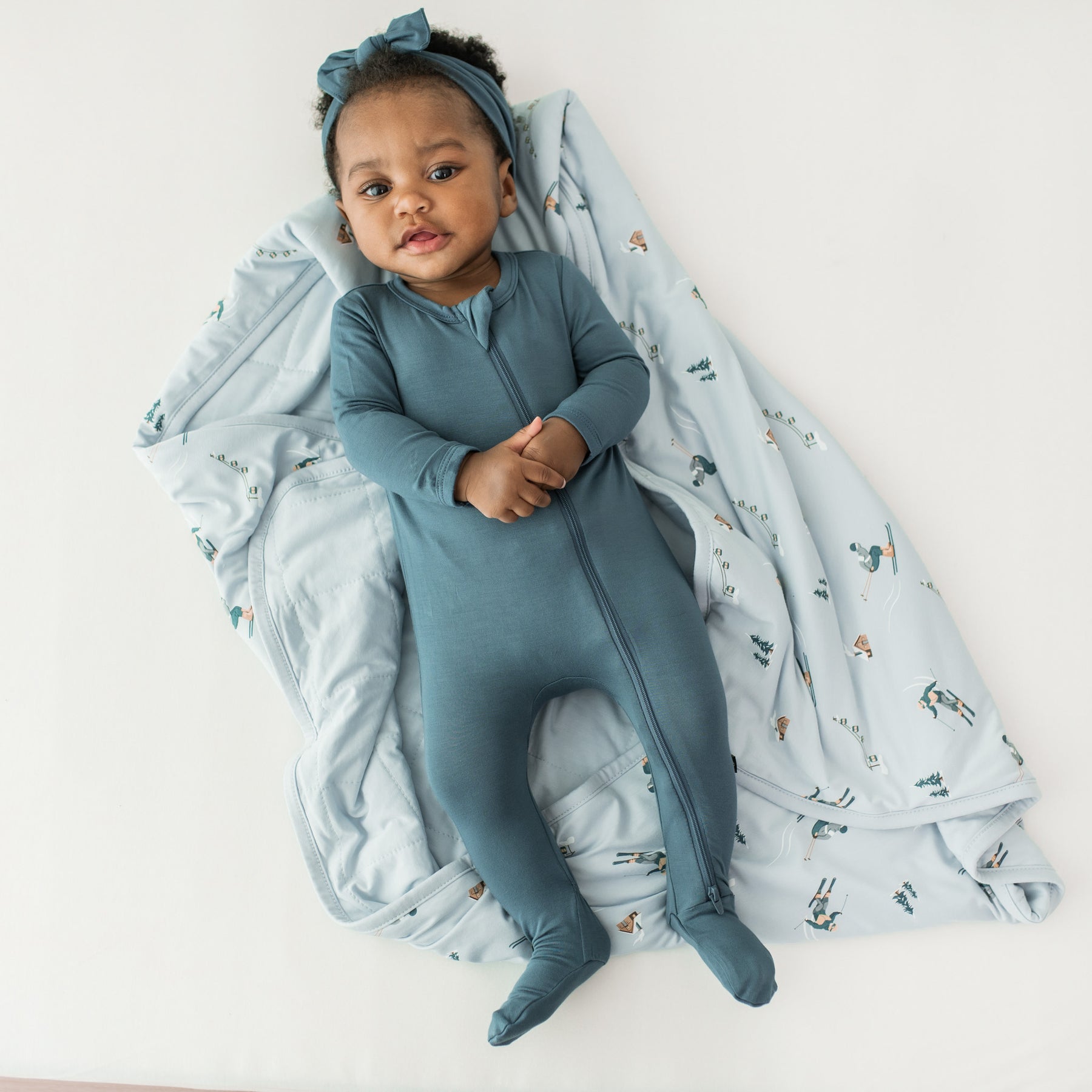 Infant girl laying on the Baby Blanket in Ski wearing a Zippered Footie in Atlantic with a matching bow headband