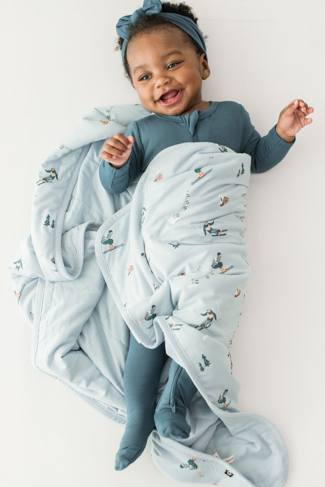 Smiling infant laying down wrapped in a Baby Blanket in Ski wearing a Zippered romper in Atlantic and matching knotted bow headband