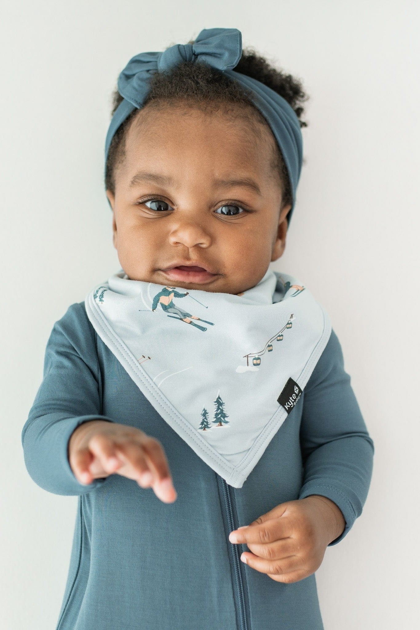 Infant laying down on a light neutral surface wearing the Bib in Ski overtop a zippered footie in Atlantic