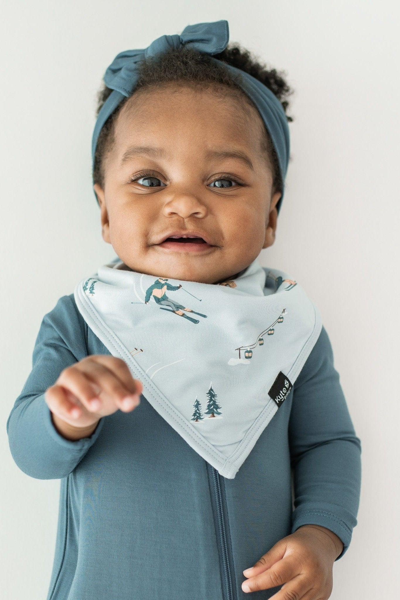 Close up of infant wearing the Bib in Ski with a zippered romper in Atlantic underneath and paired with a knotted bow headband in Atlantic