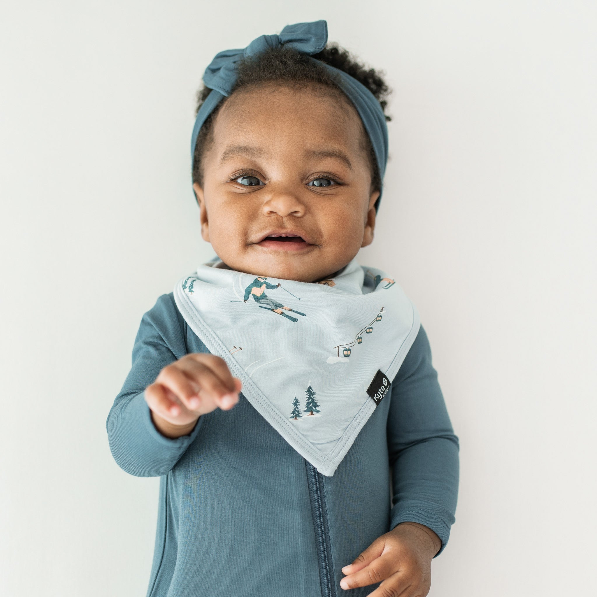 Close up of infant wearing the Bib in Ski with a zippered romper in Atlantic underneath and paired with a knotted bow headband in Atlantic