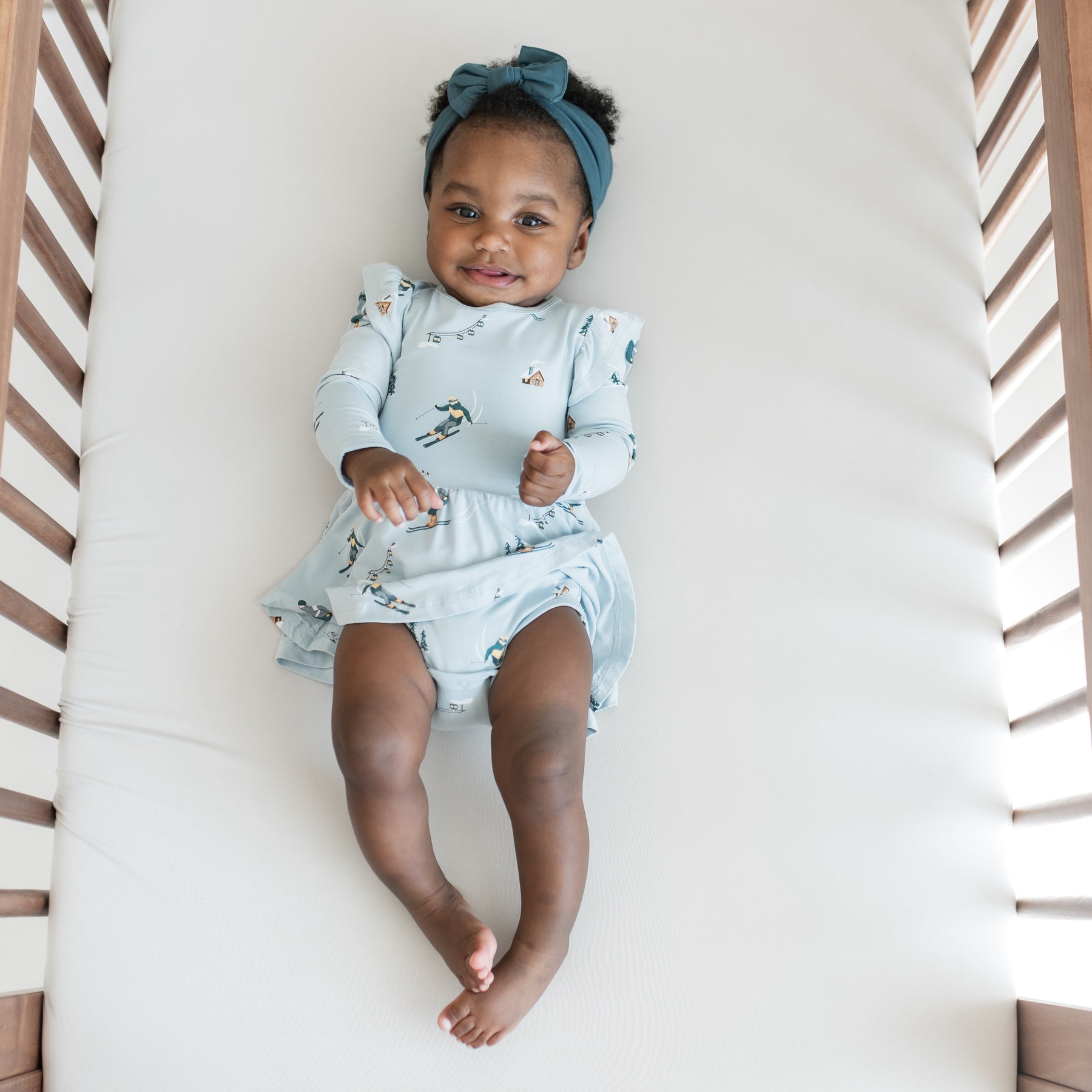 Smiling infant laying on a crib wearing the Long Sleeve Twirl Bodysuit Dress in Ski with Atlantic knotted bow headband