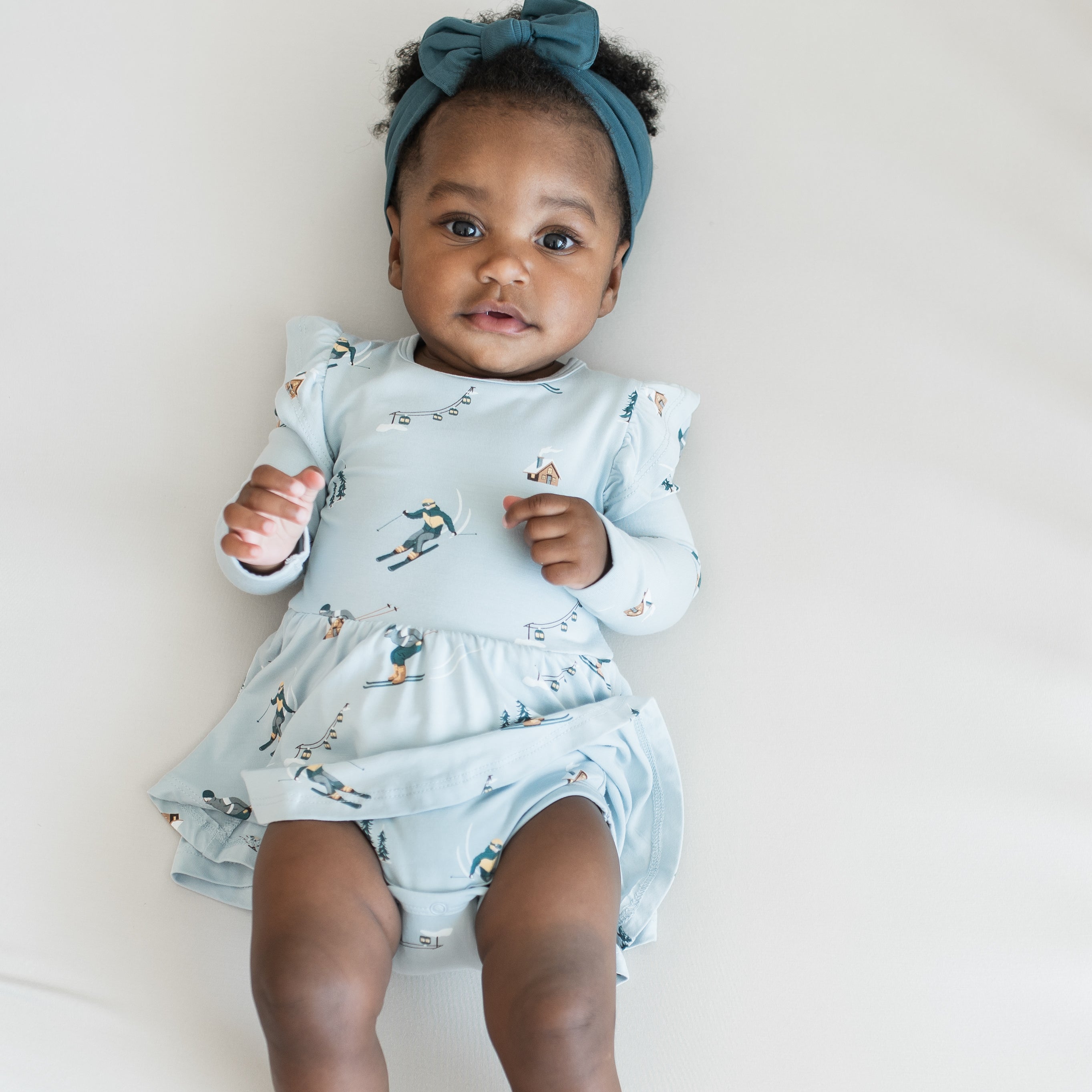 Close up of infant girl laying on a light neutral surface wearing the Long Sleeve Twirl Bodysuit Dress in Ski with Atlantic knotted bow headband 