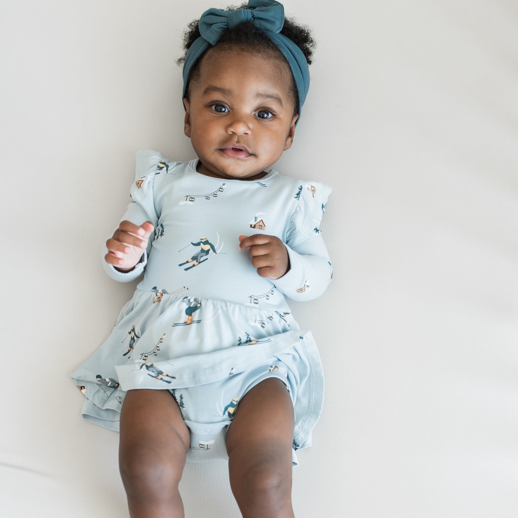 Close up of infant girl laying on a light neutral surface wearing the Long Sleeve Twirl Bodysuit Dress in Ski with Atlantic knotted bow headband 
