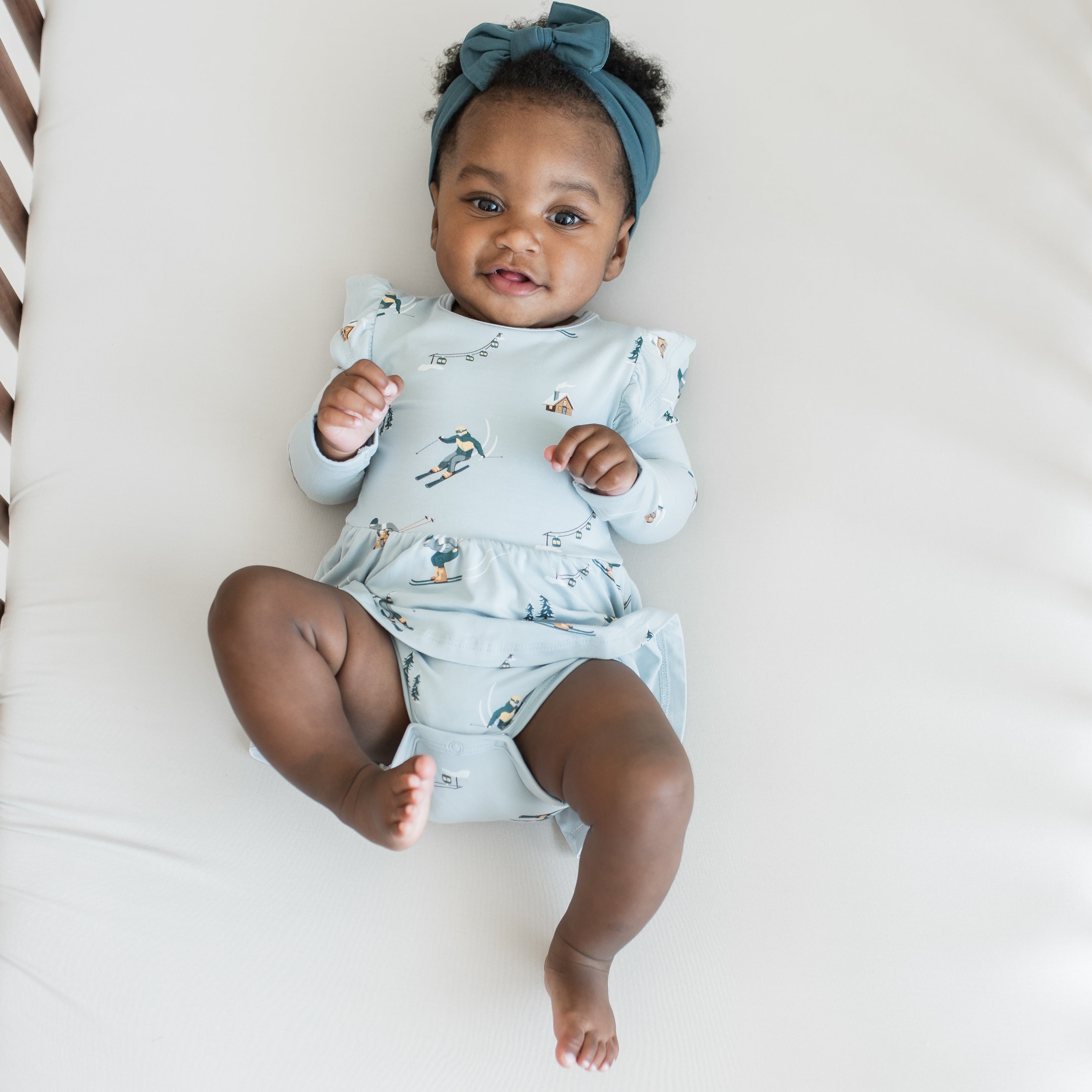 Smiling infant laying in a crib on a light neutral crib sheet wearing the Long Sleeve Twirl Bodysuit Dress in Ski