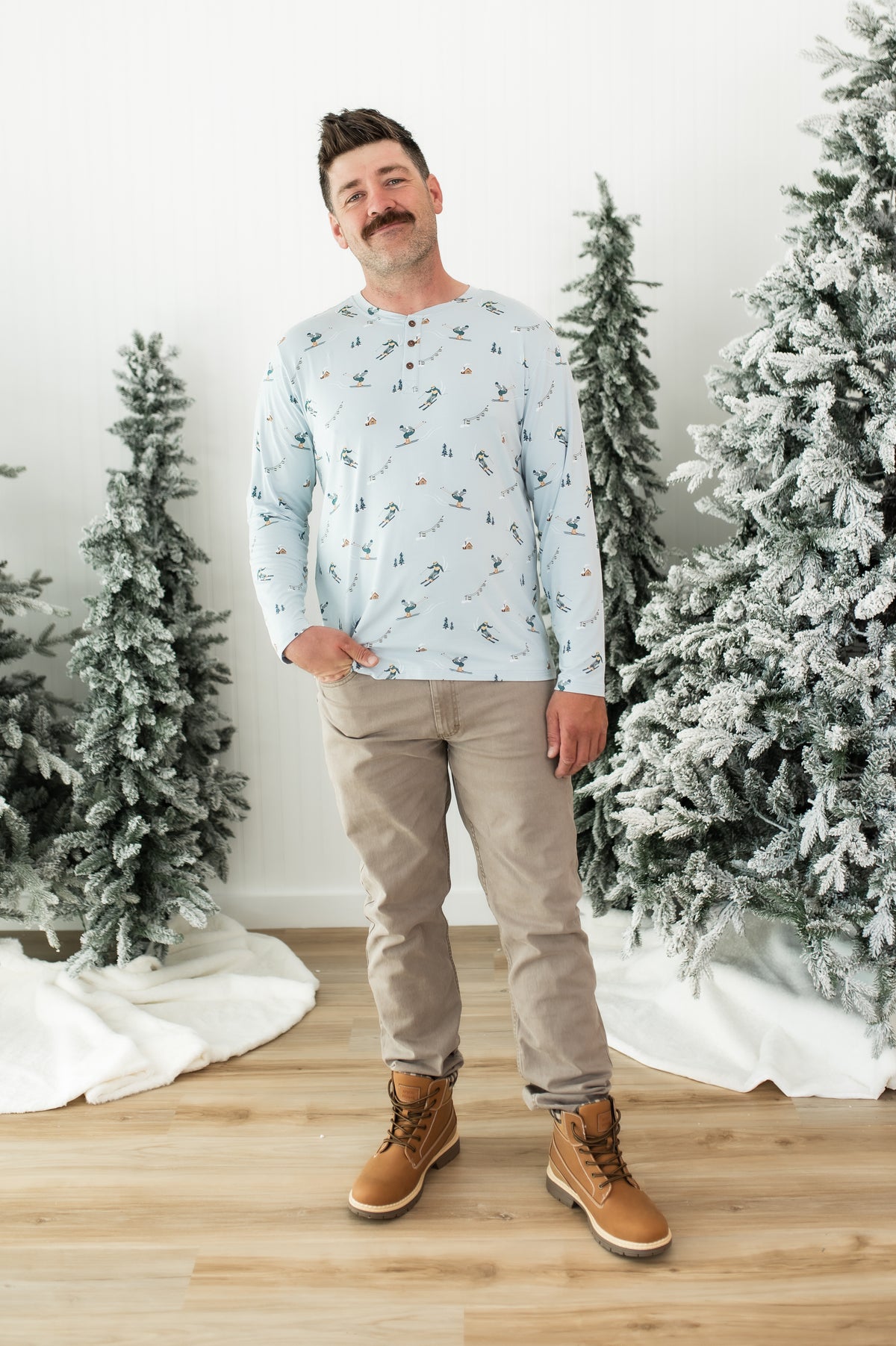 Smiling male modeling the Men's Long Sleeve Henley Top in Ski with khaki colored pants and light brown boots standing between frosted trees