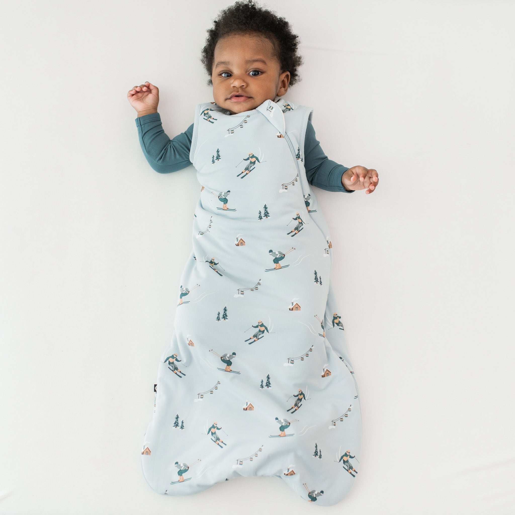 Young toddler laying on a light neutral surface wearing the Sleep Bag in Ski 1.0 with a muted blue romper underneath