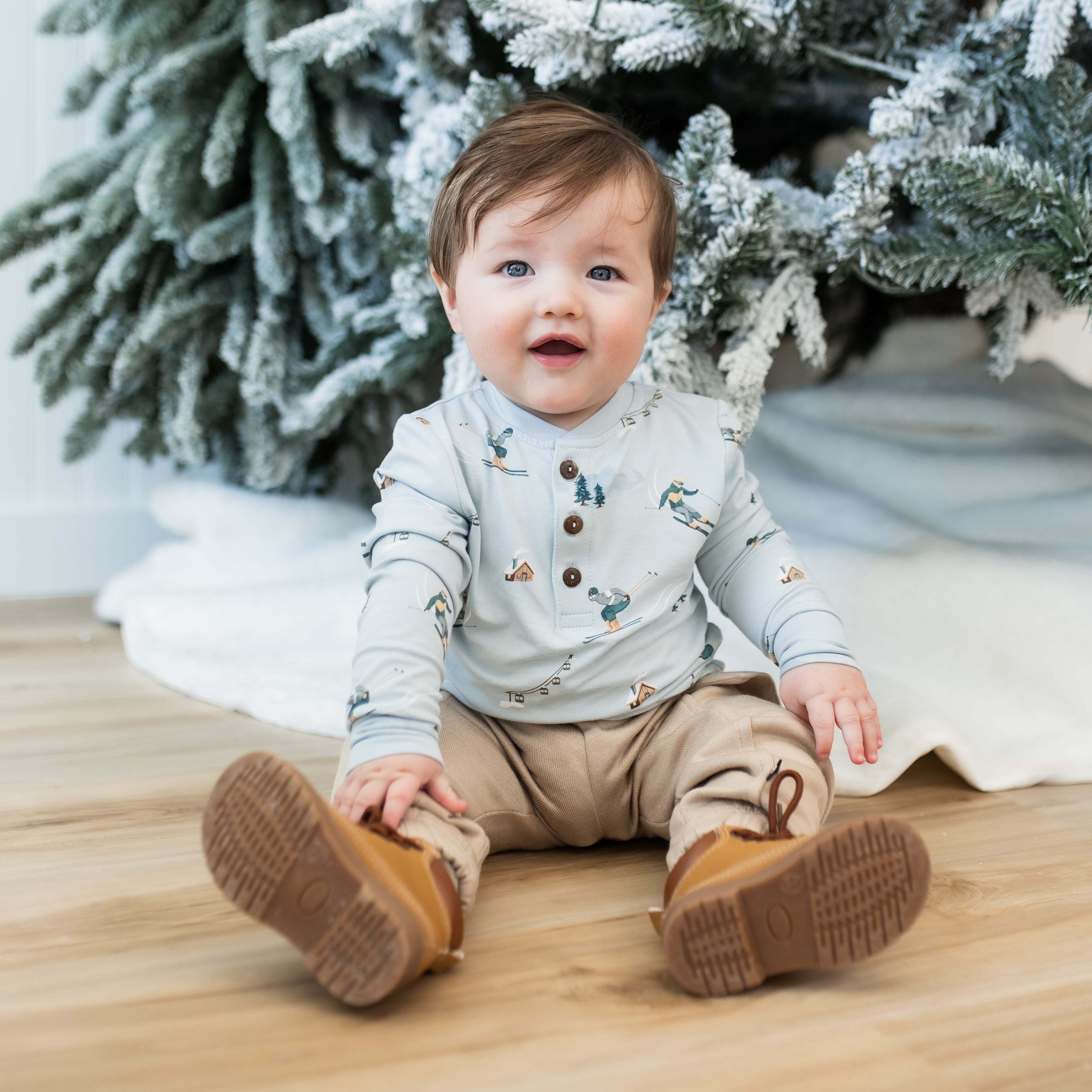 Toddler sitting on the floor in front of a frosted tree wearing the Long Sleeve Toddler Henley Tee in Ski with light tan pants