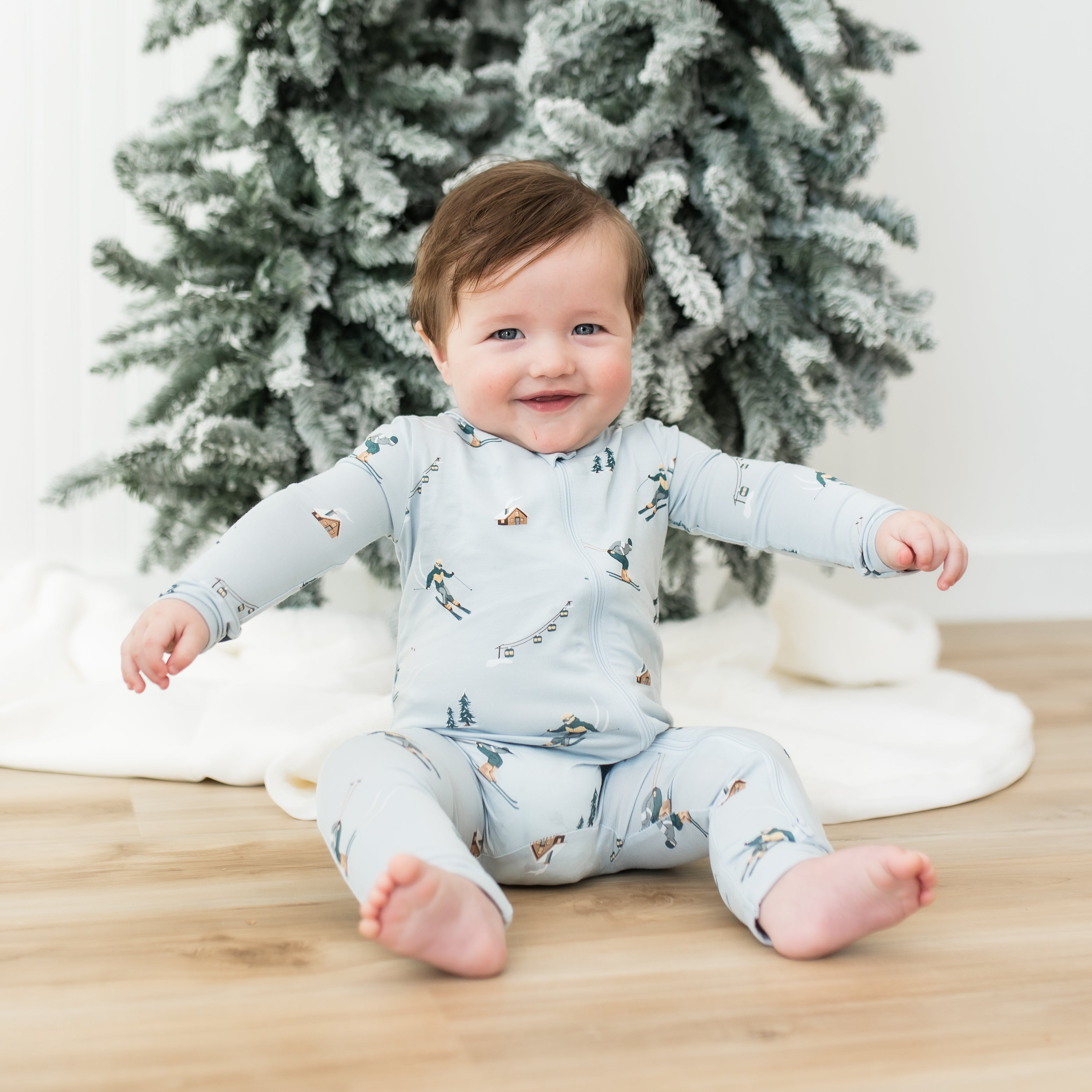 Young toddler sitting on the floor in front of a frosted tree wearing the Zippered Romper in Ski
