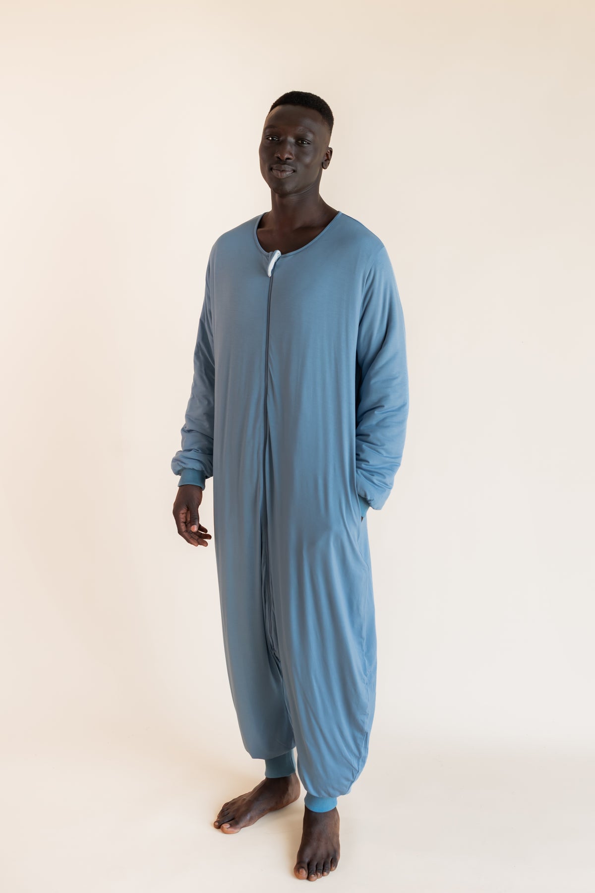A man wearing a dusty blue long-sleeve zip-up onesie, standing against a plain beige studio background.