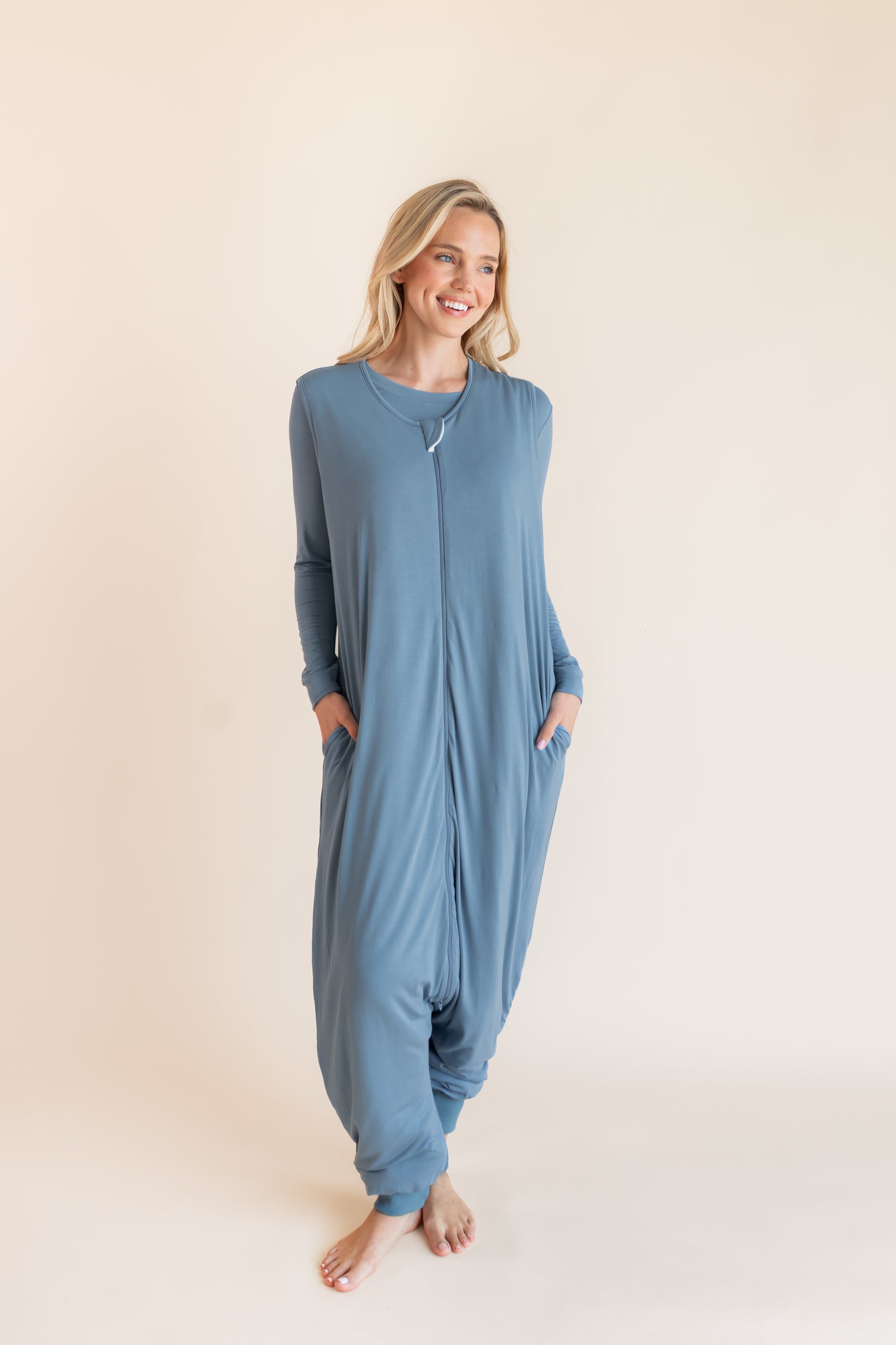 A woman wearing a dusty blue sleep sack pajama one-piece with a front zipper and side pockets, standing against a solid beige studio background.