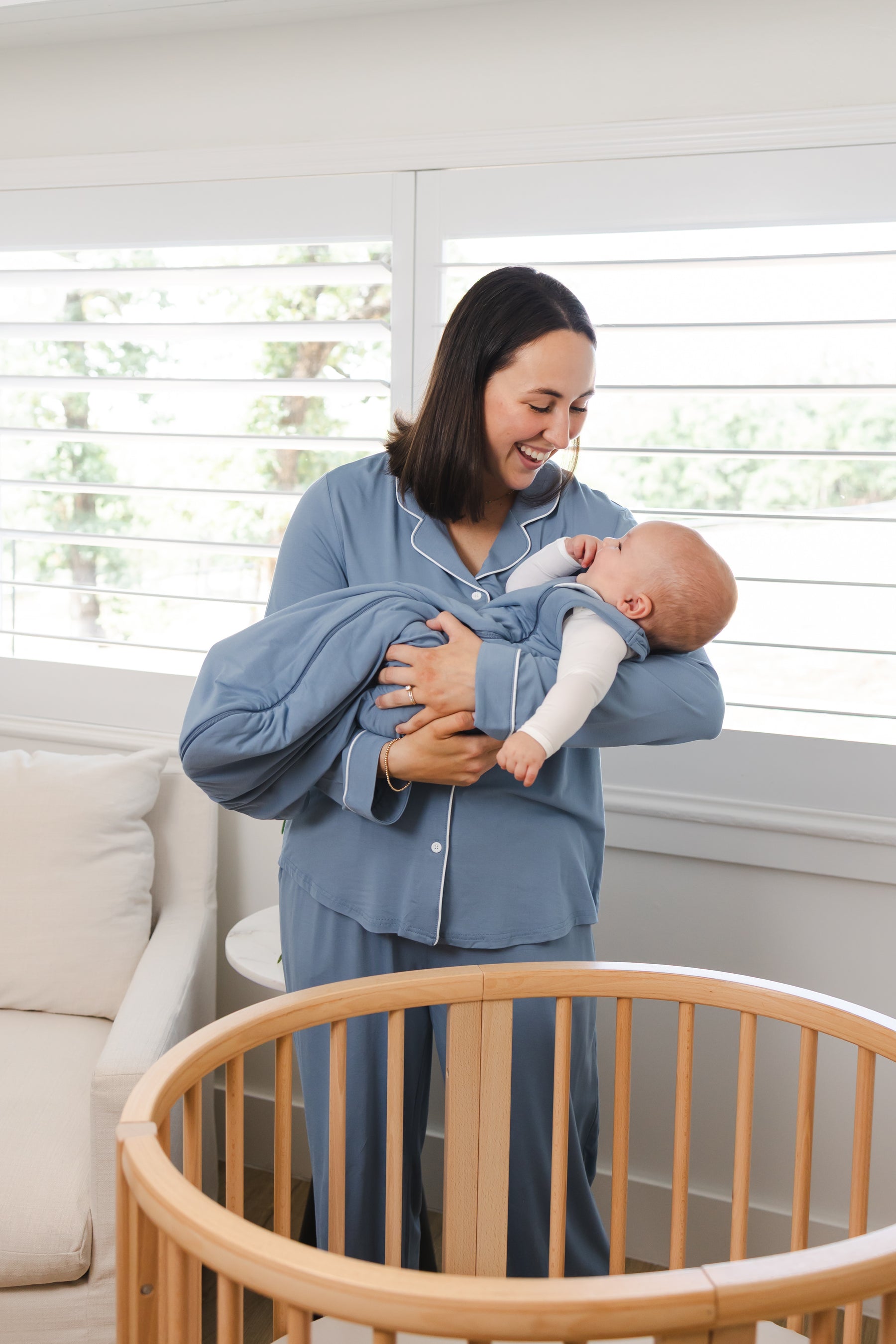 A woman in dusty blue pajamas holds a baby wrapped in a matching blue sleep sack in a bright nursery with white shutters and a wooden crib.