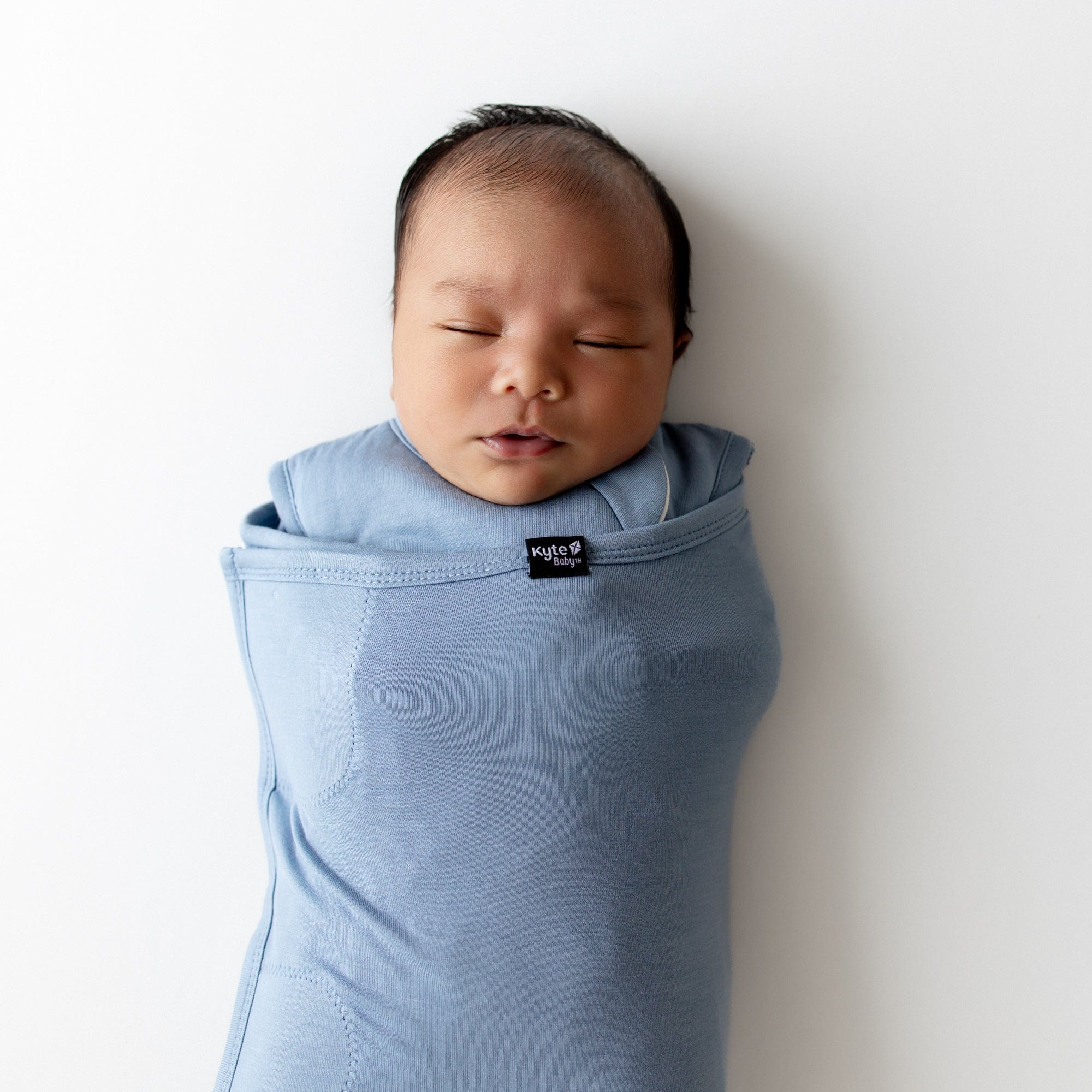 Newborn baby wrapped in a blue swaddler with a Kyte Baby label on a white background