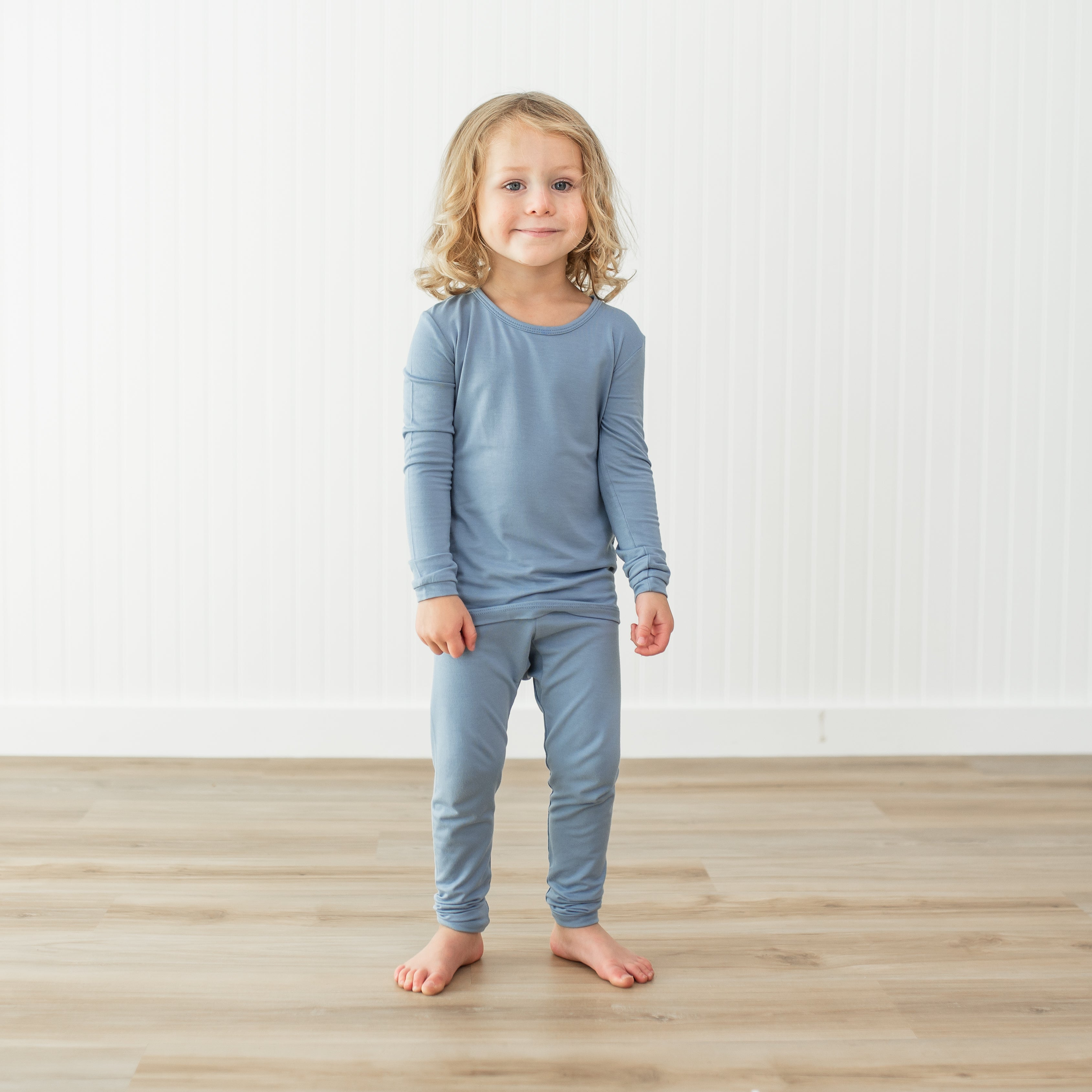 Child wearing a blue pajama set standing on a wooden floor with a white wall background