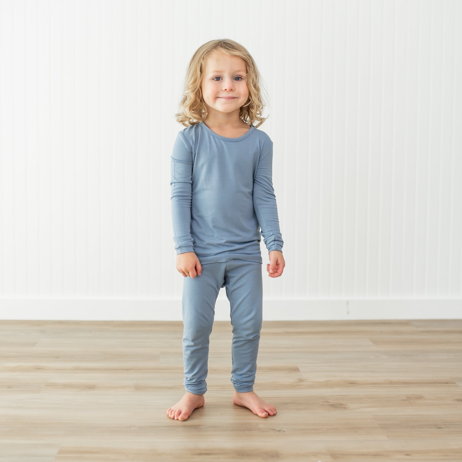 Child wearing a blue pajama set standing on a wooden floor with a white wall background