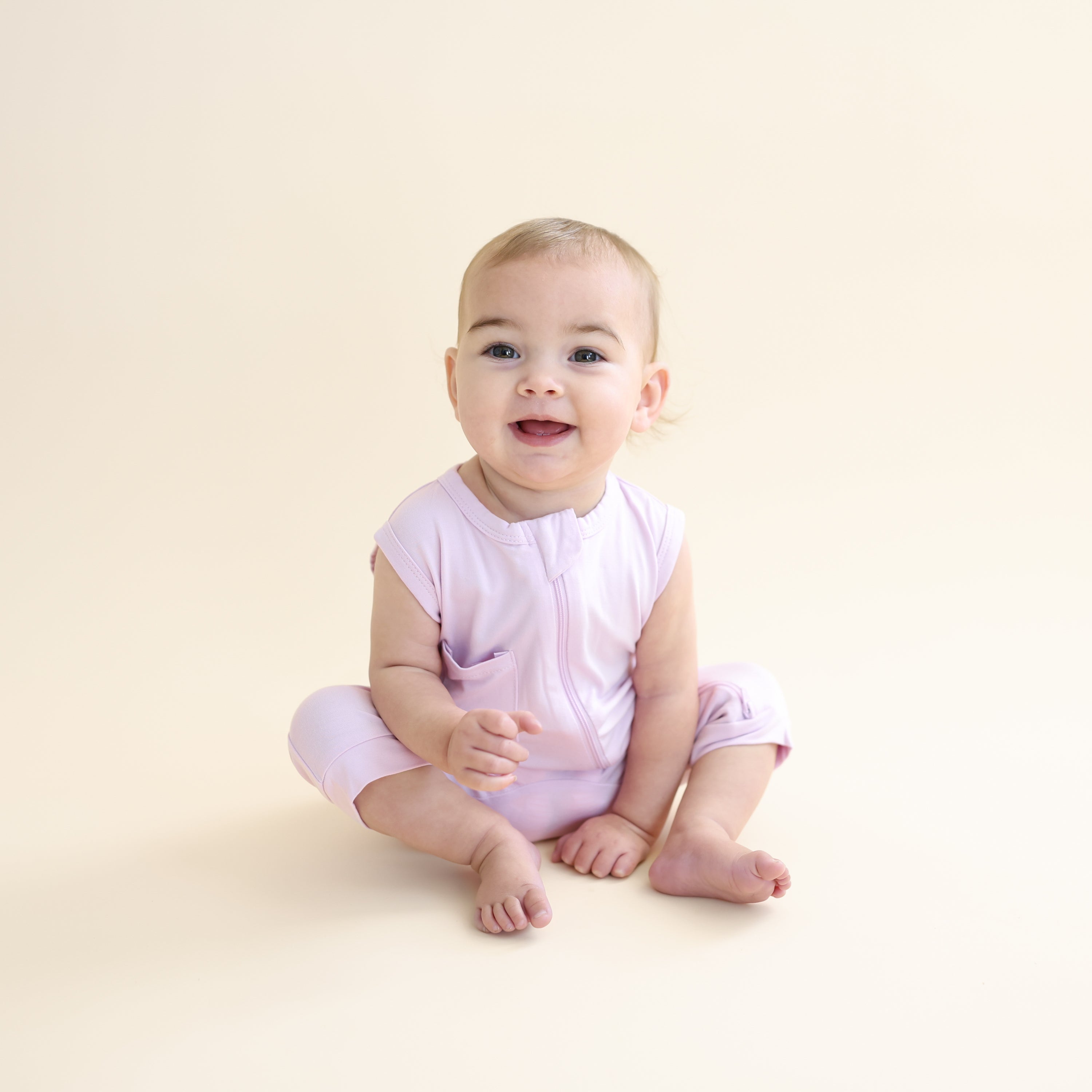 Smiling toddler sitting on the floor wearing the Zippered Sleeveless Romper in Thistle