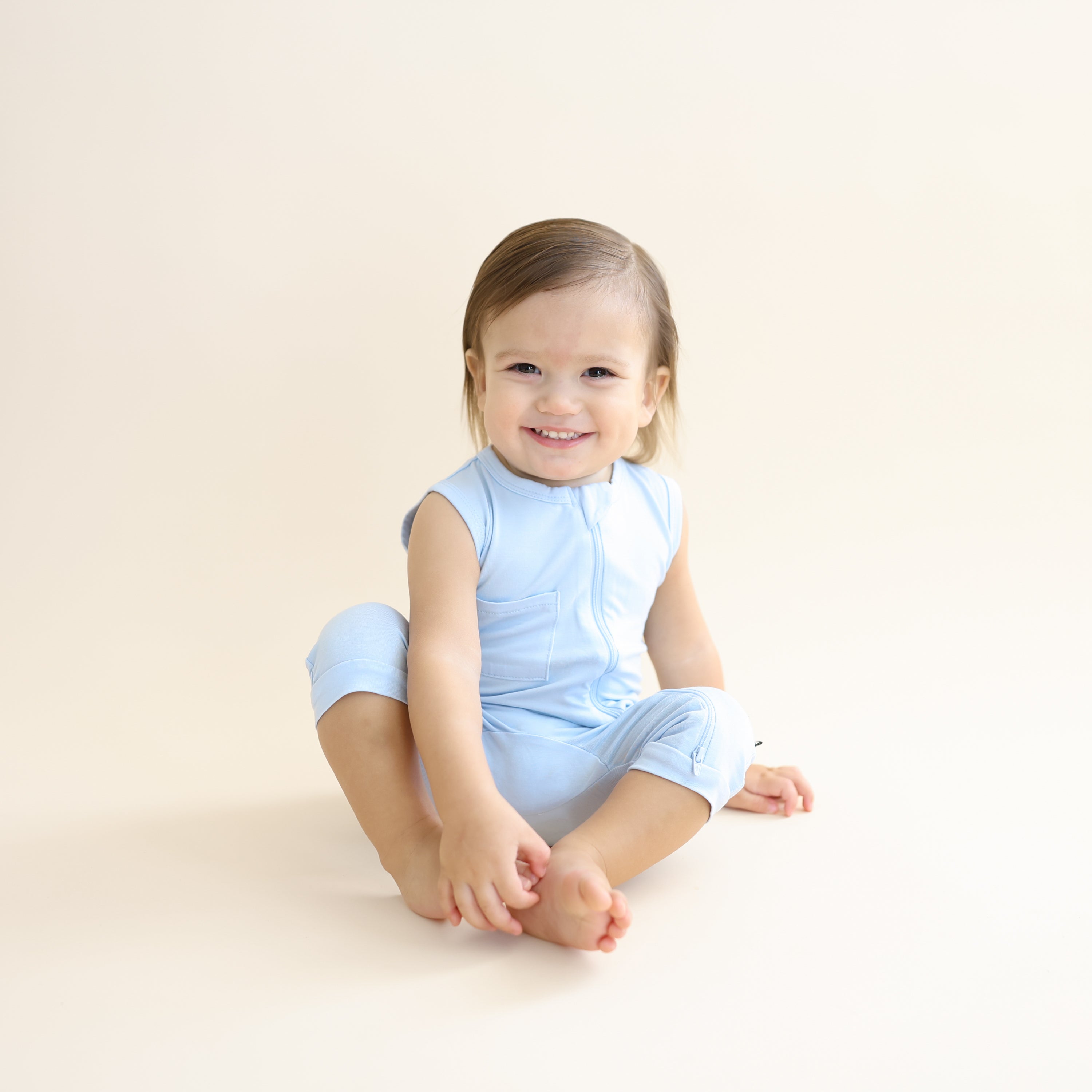 Smiling toddler sitting on the floor wearing the Zippered Sleeveless Romper in Breeze