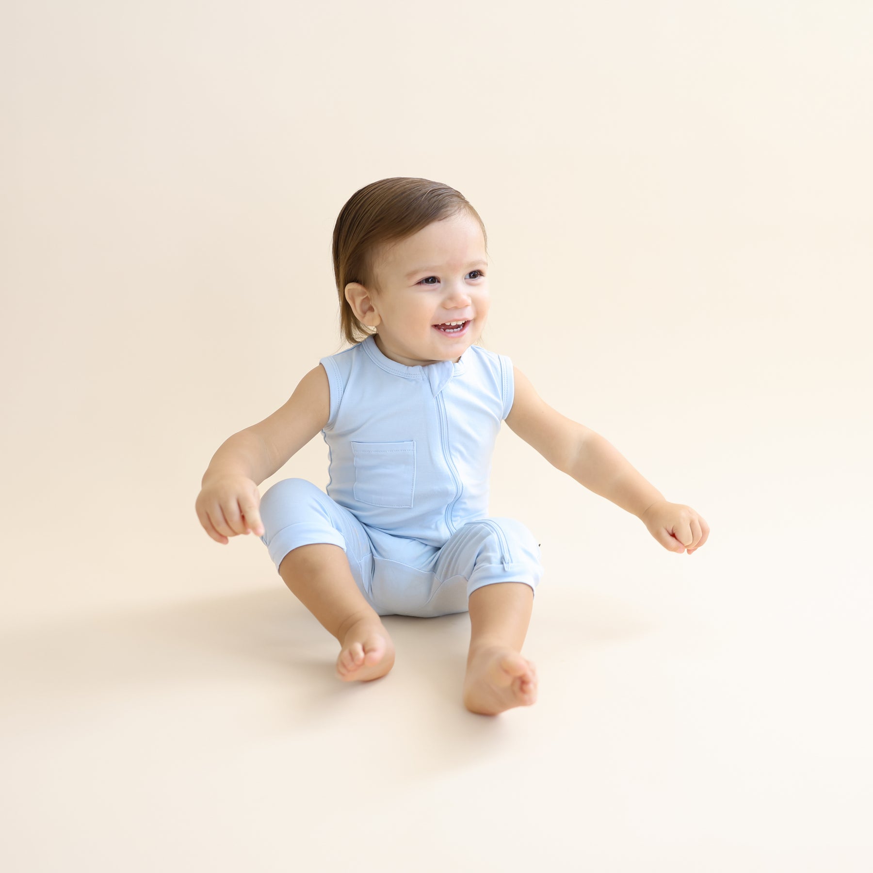 Young toddler sitting on the floor wearing the Zippered Sleeveless Romper in Breeze