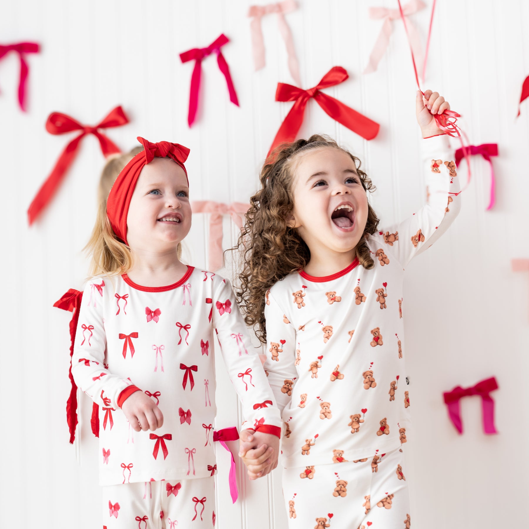 Two girls standing in front of a white paneled wall with bows on it wearing the Long Sleeve Pajamas in Small Love Bow and Bear Hearts