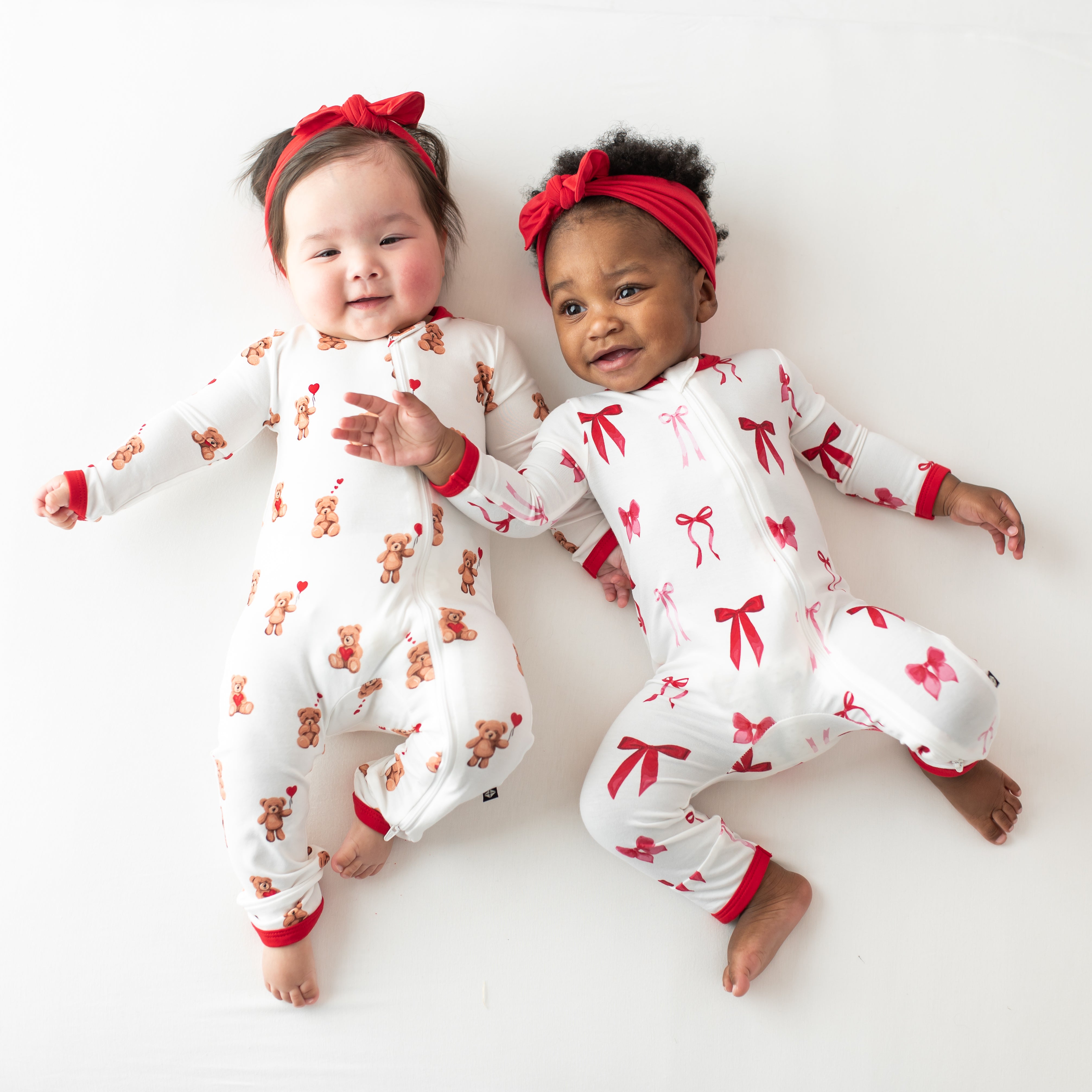 Two young toddlers laying side by side wearing the Zippered Romper in Small Love Bow and Bear Hearts with Red bows