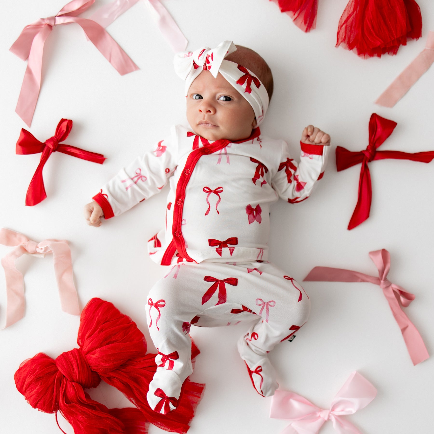 Newborn laying on a white surface wearing the soft and breathable Take Me Home Set with Bow in Small Love Bow surrounded by various types of bows