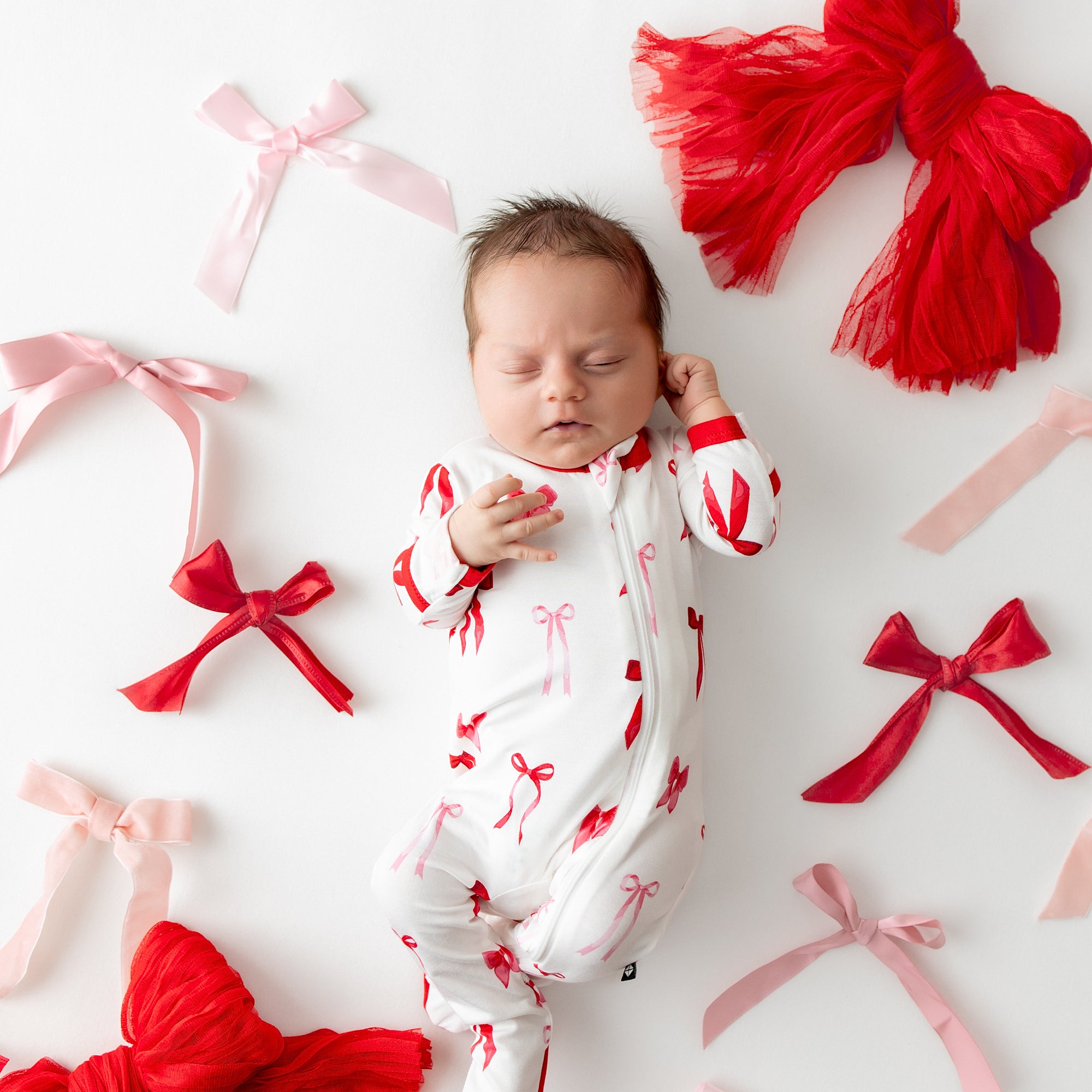 Sleeping infant wearing the Zippered Footie in Small Love Bow surrounded by various bows